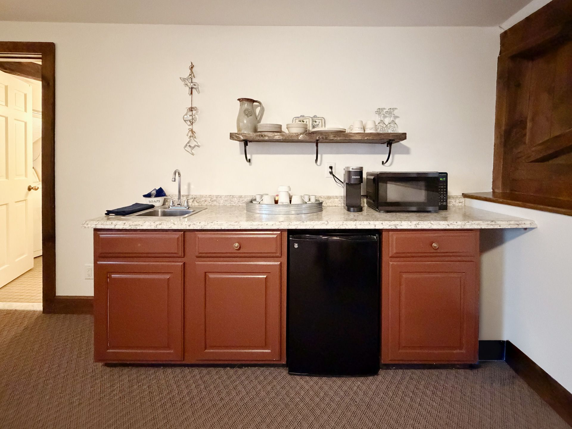 Small kitchen counter with sink, cabinets, mini fridge, microwave, and shelf with dishes