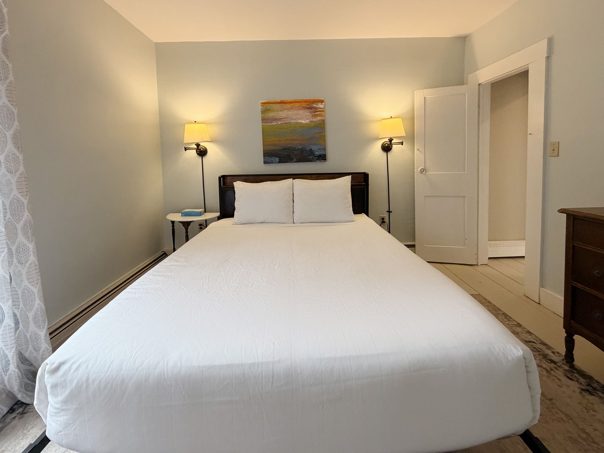 Neatly made bed in a bright bedroom with two lamps and a small painting above the headboard