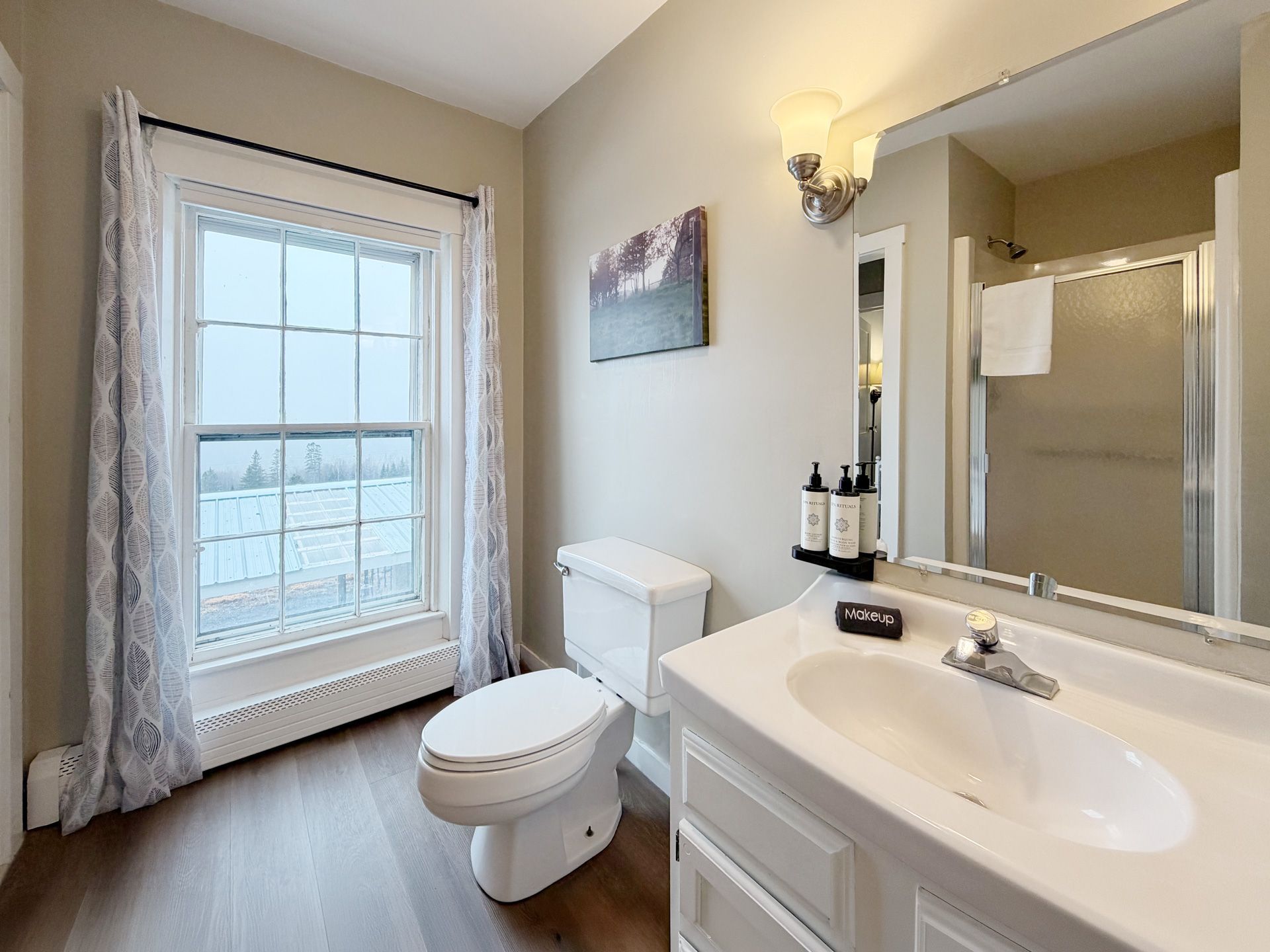 Bright bathroom with toilet, sink, mirror, walk-in shower, and large window with curtain.