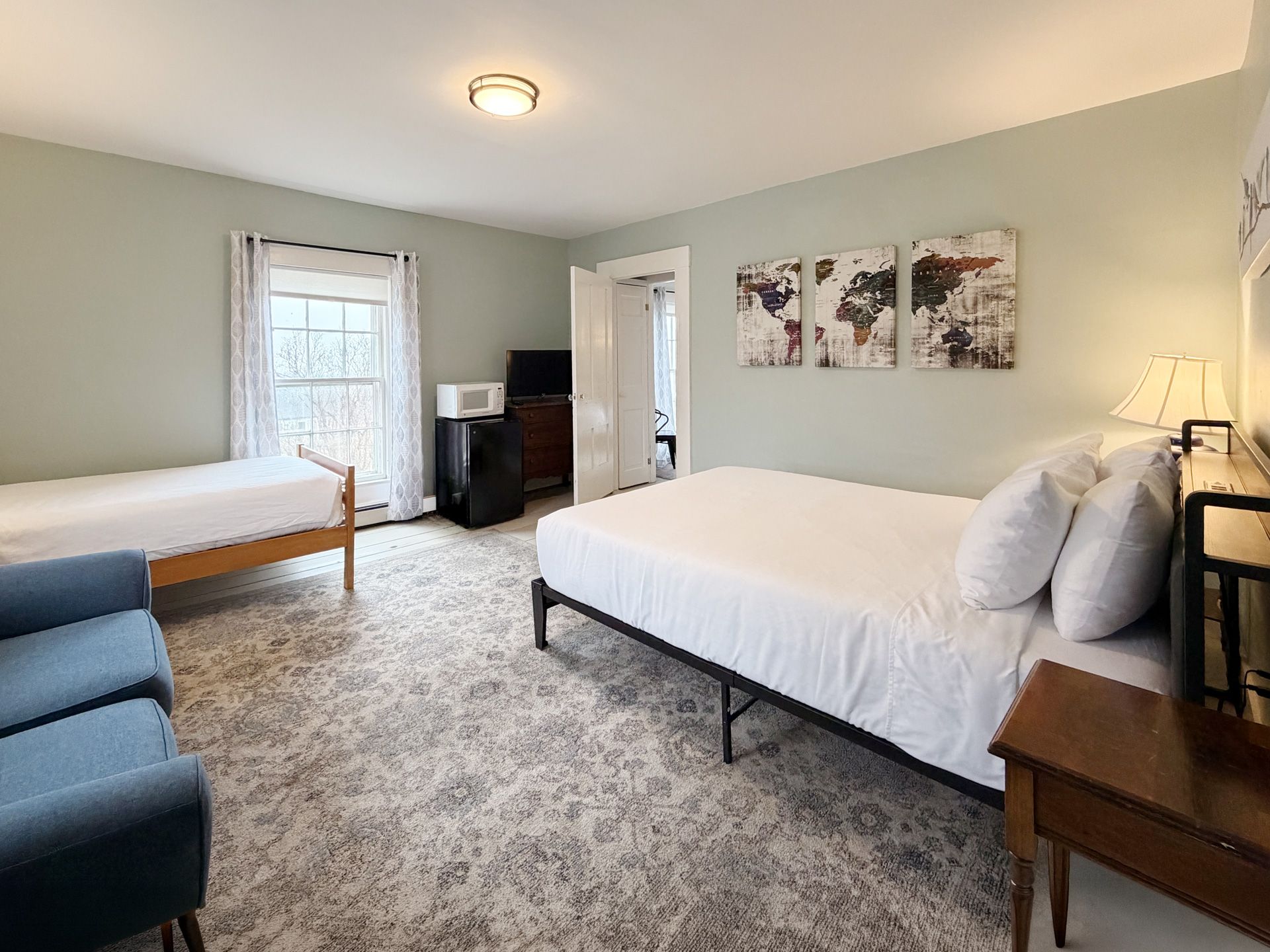 Bright bedroom with two beds, blue chair, white walls, and a dresser near a window and lamp.