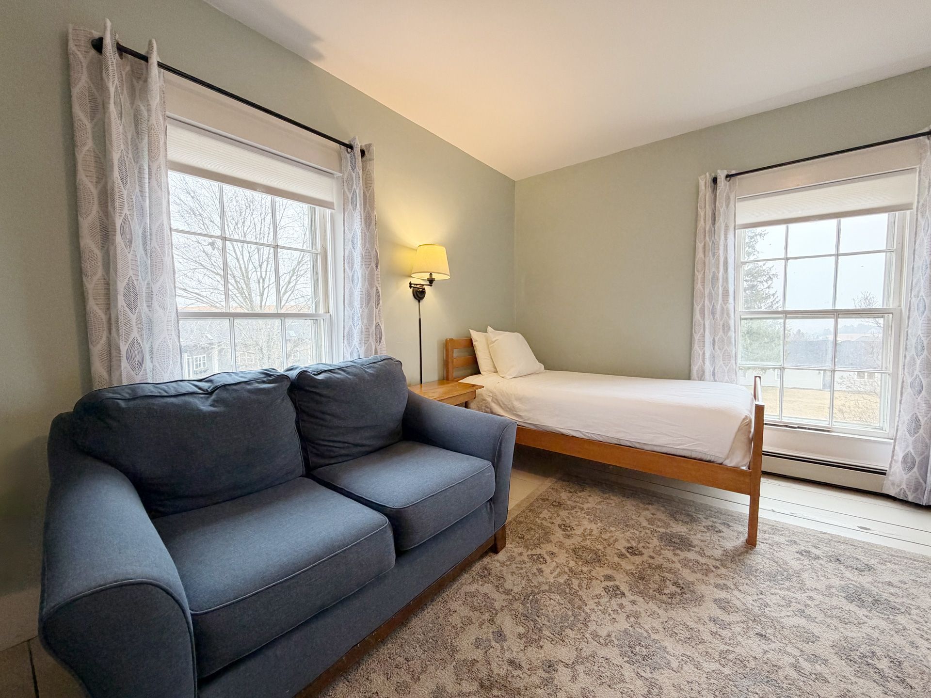 Bright bedroom with a blue sofa, bed, two windows, and a floor lamp on a patterned rug.