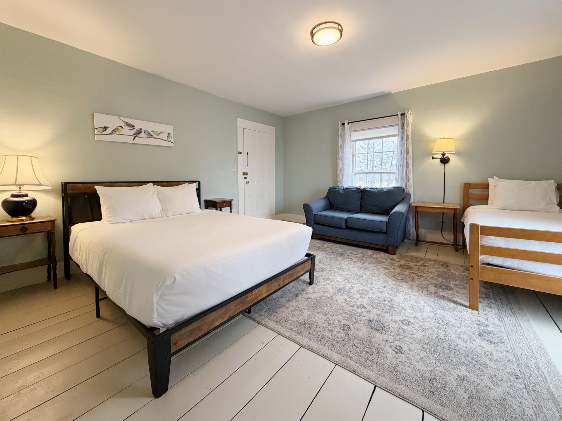 Bright bedroom with queen bed, blue loveseat, desk, and window, in soft gray tones