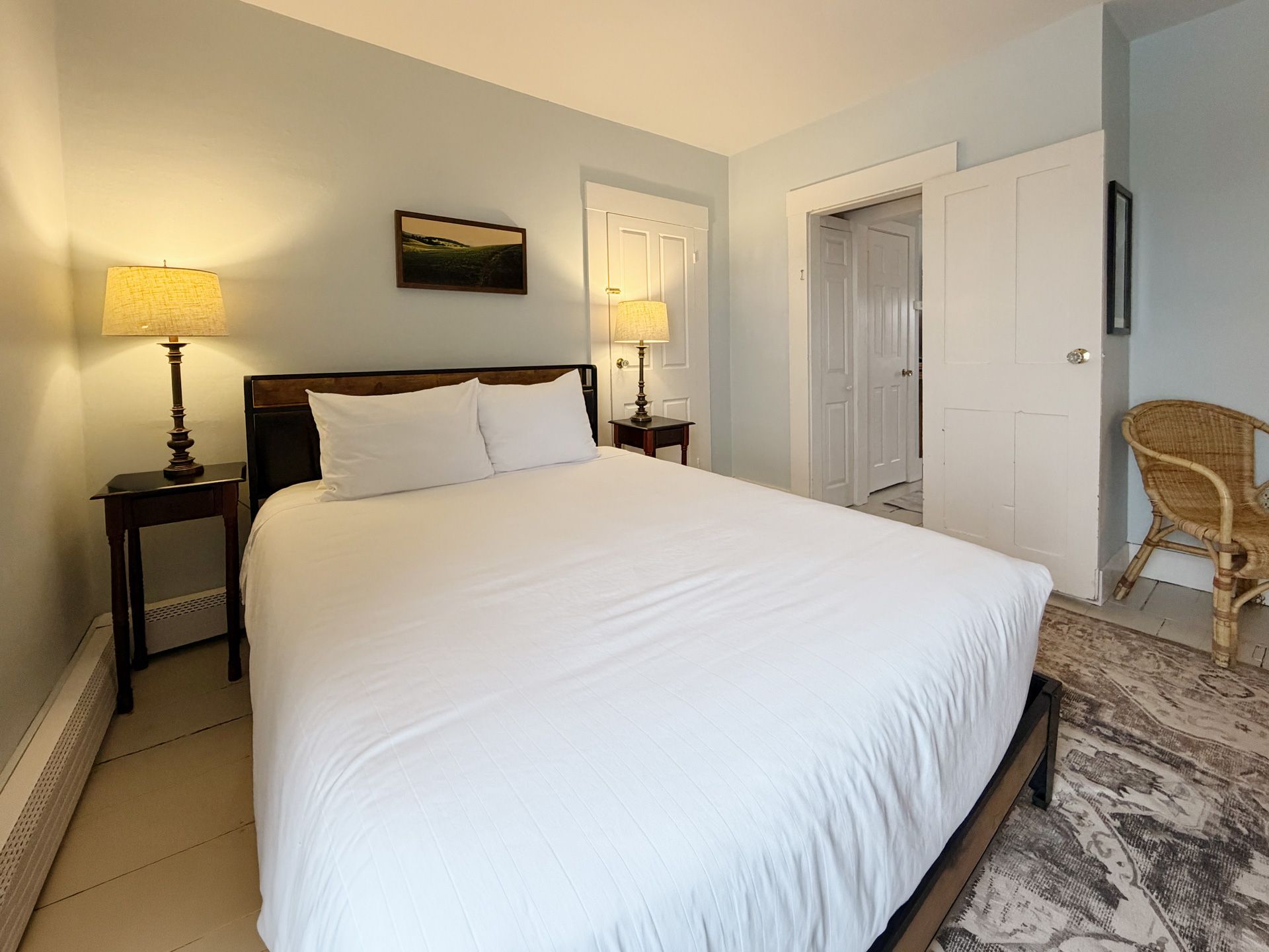 Bedroom with white bedding, two bedside lamps, a closet, and wicker chair on carpeted floor.