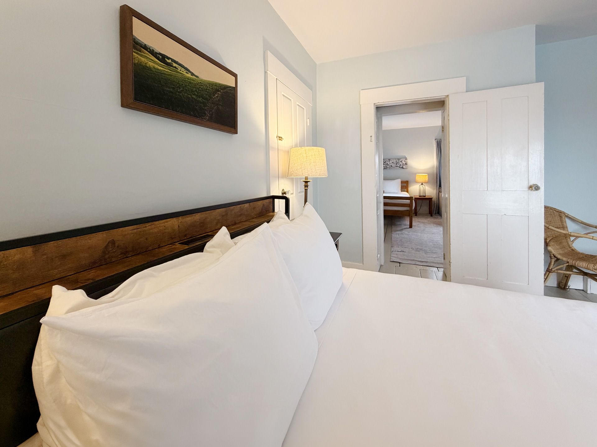 Bright bedroom with white bedding, wooden headboard, lamp, and open door to a hallway and adjoining room.