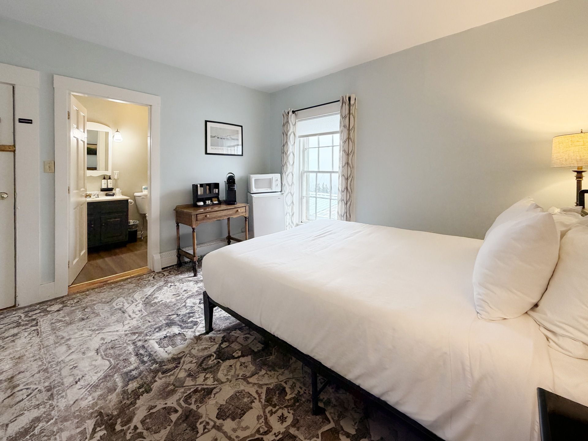 Bright bedroom with a made white bed, patterned rug, dresser, and open doorway to a bathroom