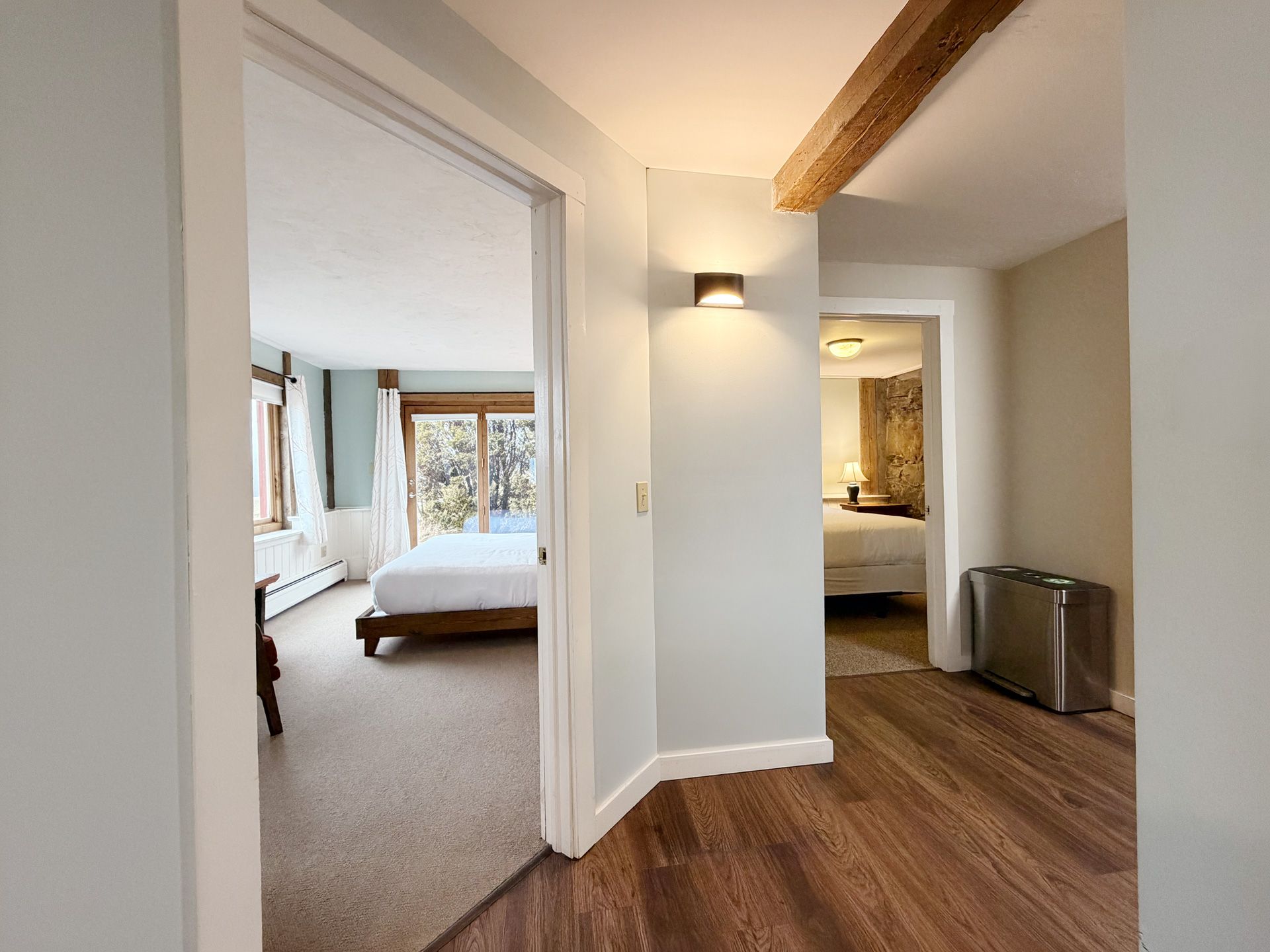 Bright apartment hallway with wood floor, wall sconce, and open doorways to carpeted bedrooms.