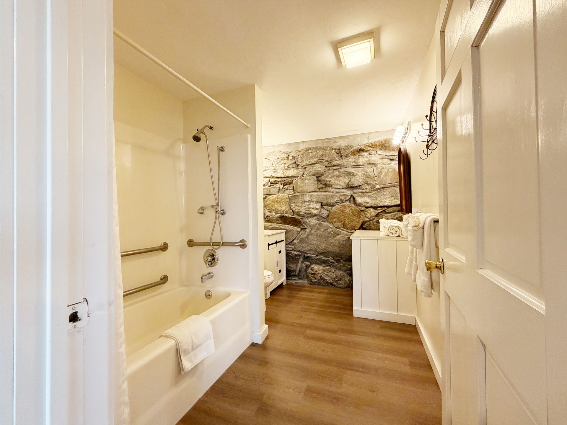 Narrow bathroom with white tub, shower fixtures, wood floor, and exposed stone wall end