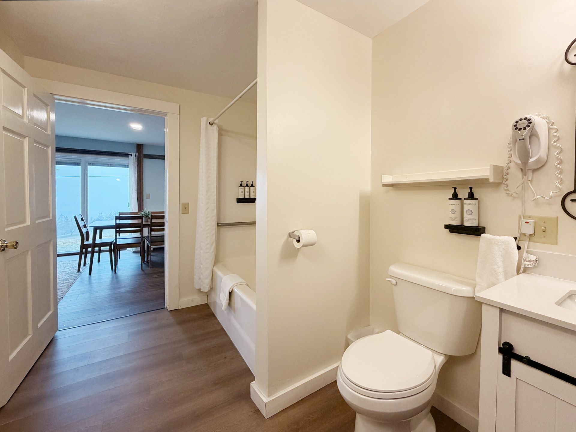 Bathroom with white toilet, sink, tub, and open door to a dining room beyond.
