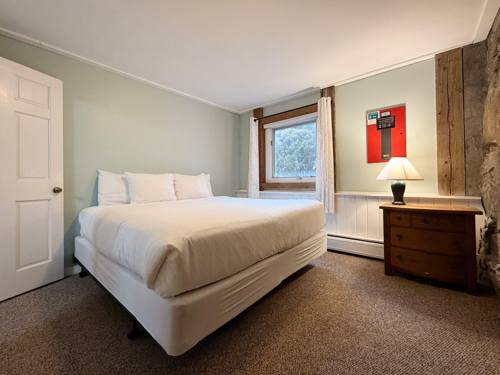 Neatly made white bed in a small bedroom with a window, dresser, lamp, and red wall art.