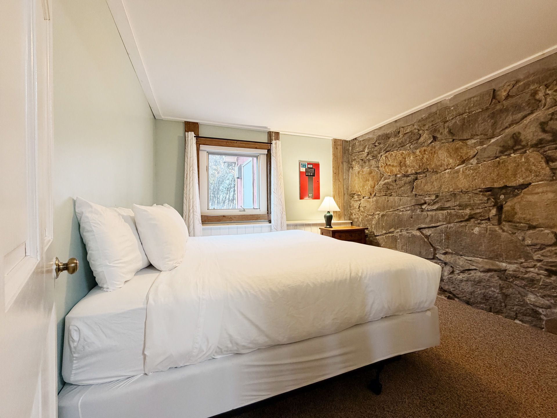 Small bedroom with a white bed, exposed stone wall, and a window letting in daylight