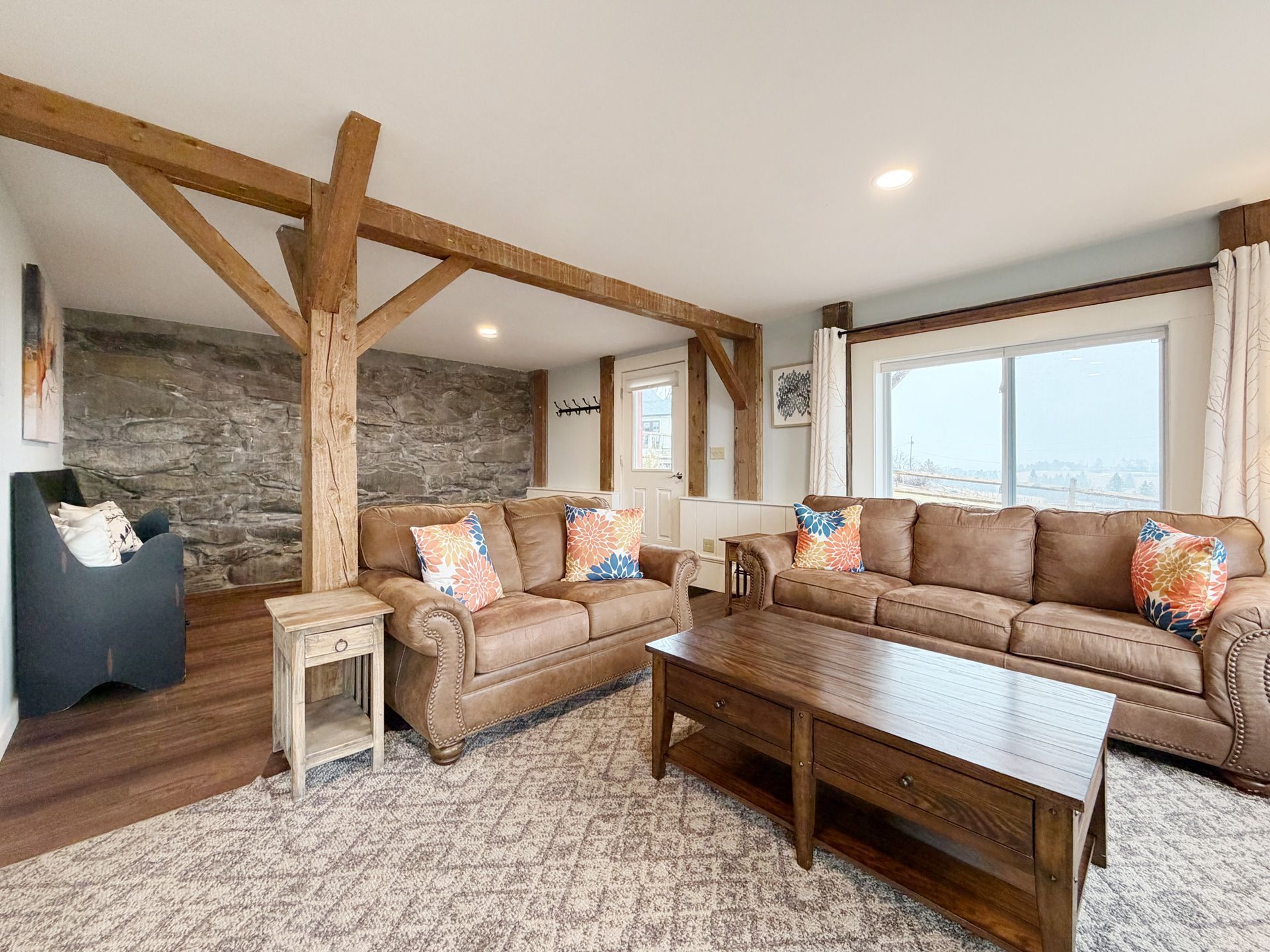 Cozy living room with tan sofas, exposed wood beams, stone fireplace, and large windows.
