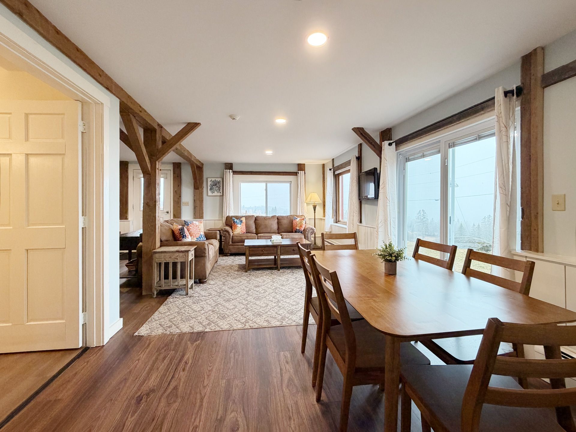 Bright open living and dining room with wood floors, white walls, exposed beams, and large windows