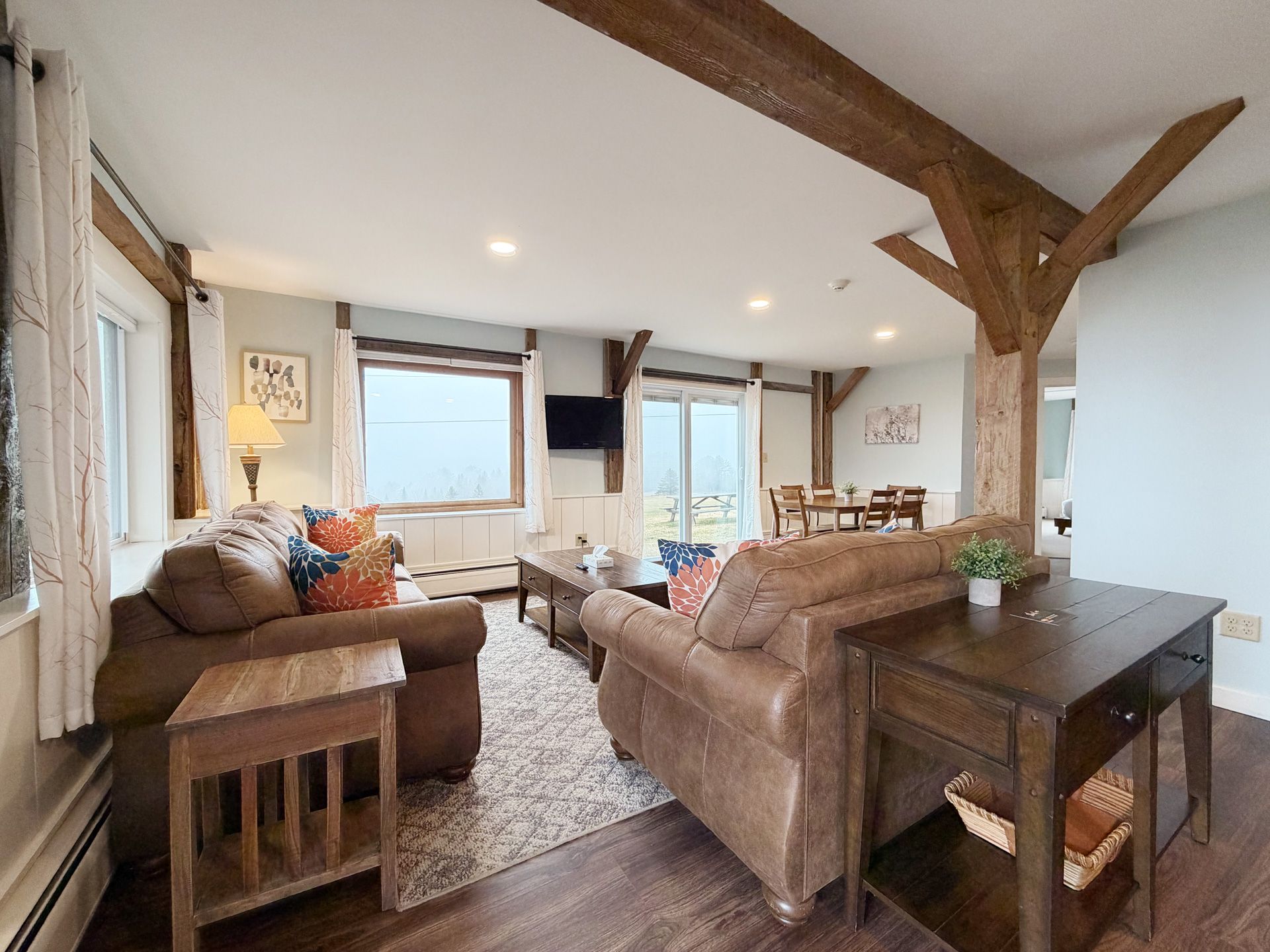 Cozy open-plan living room with brown sofas, wood beams, and large windows overlooking water