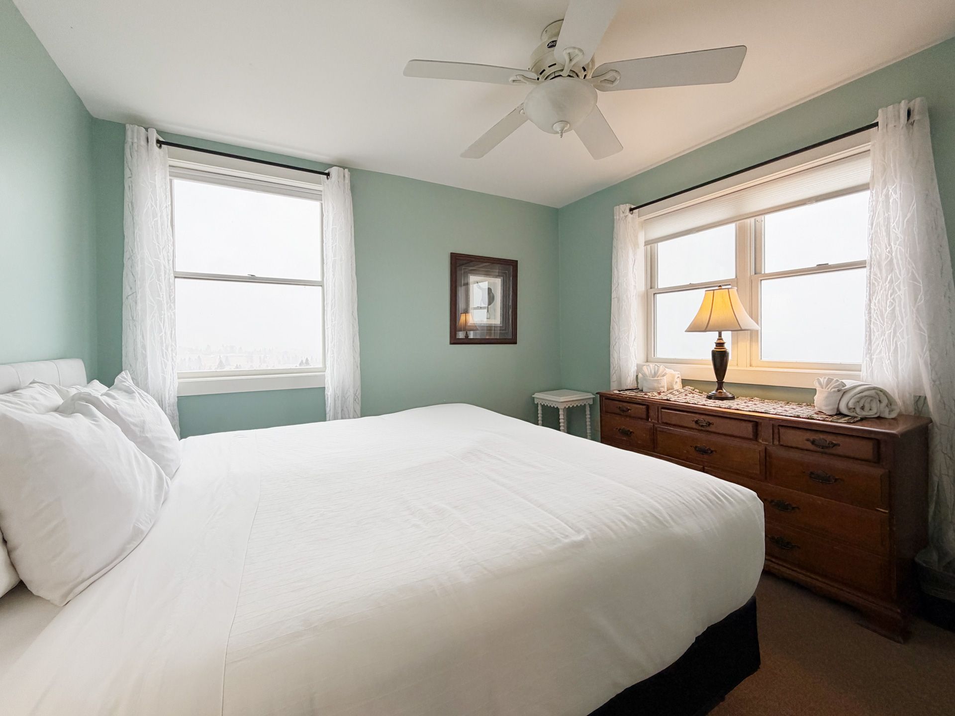 Bright bedroom with white bed, mint walls, ceiling fan, and wooden dresser by windows.