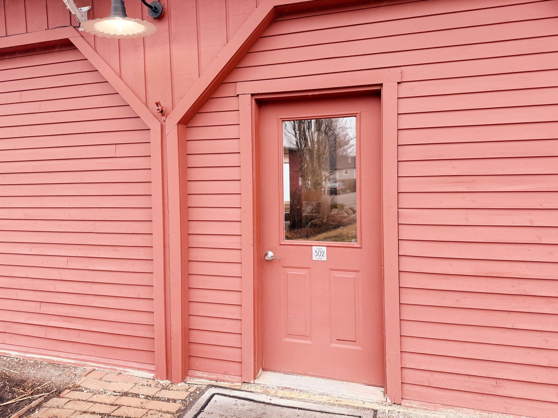 Pink wooden exterior door with a small glass pane and wall-mounted light on a matching siding wall