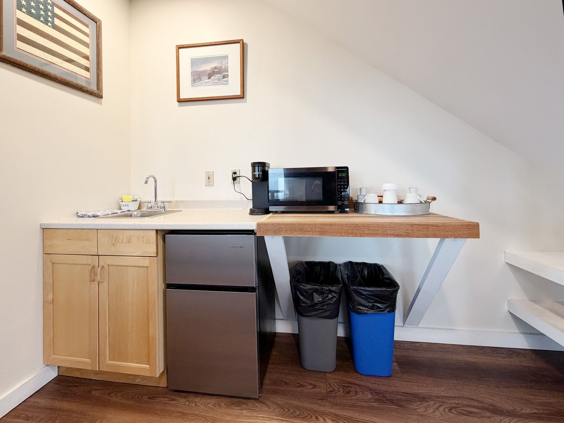 Small kitchenette with sink, mini fridge, microwave, trash bins, and wooden counter in a sloped-ceiling room