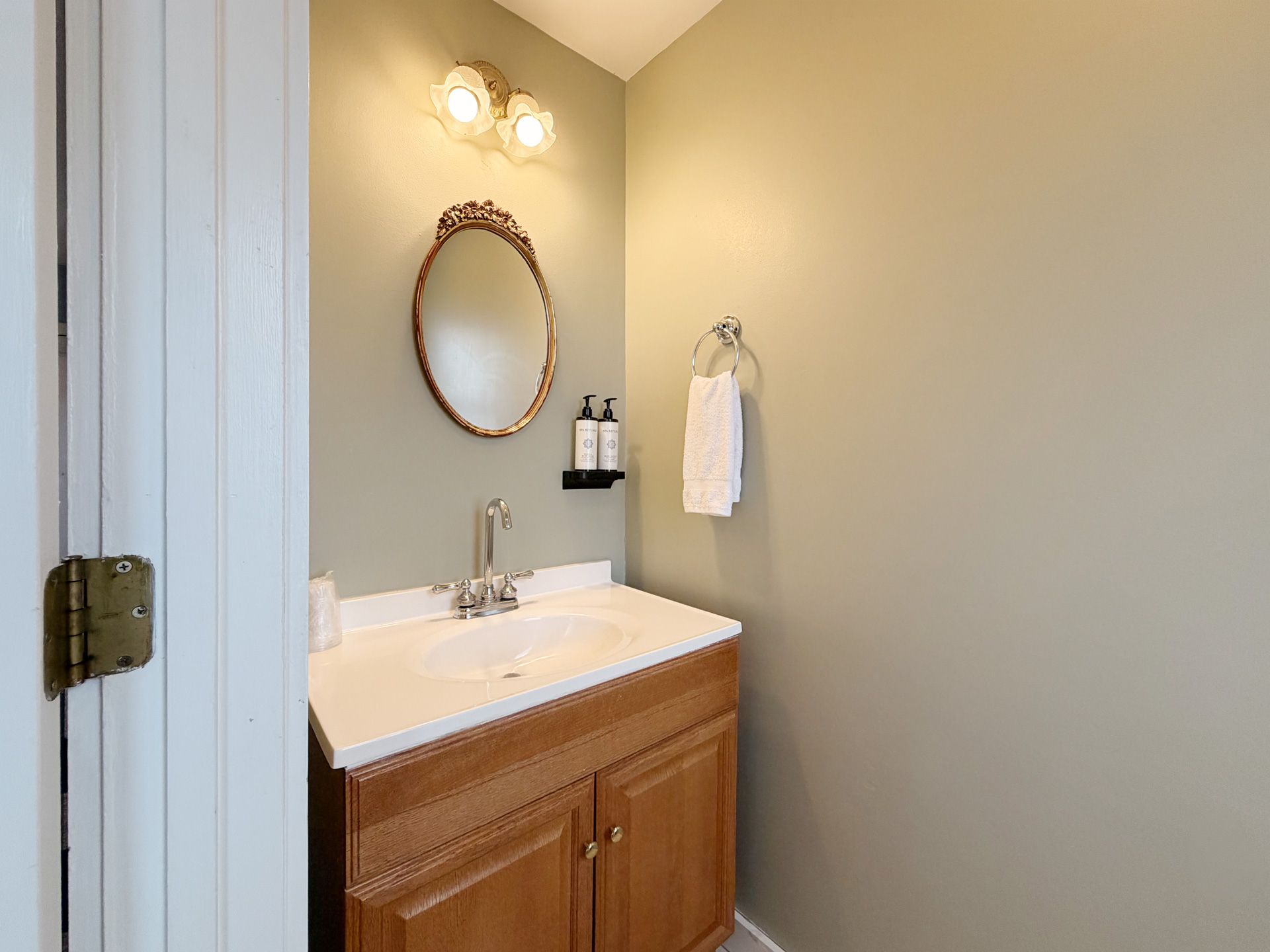 Small bathroom vanity with white sink, wood cabinet, oval mirror, and towel ring on a beige wall.