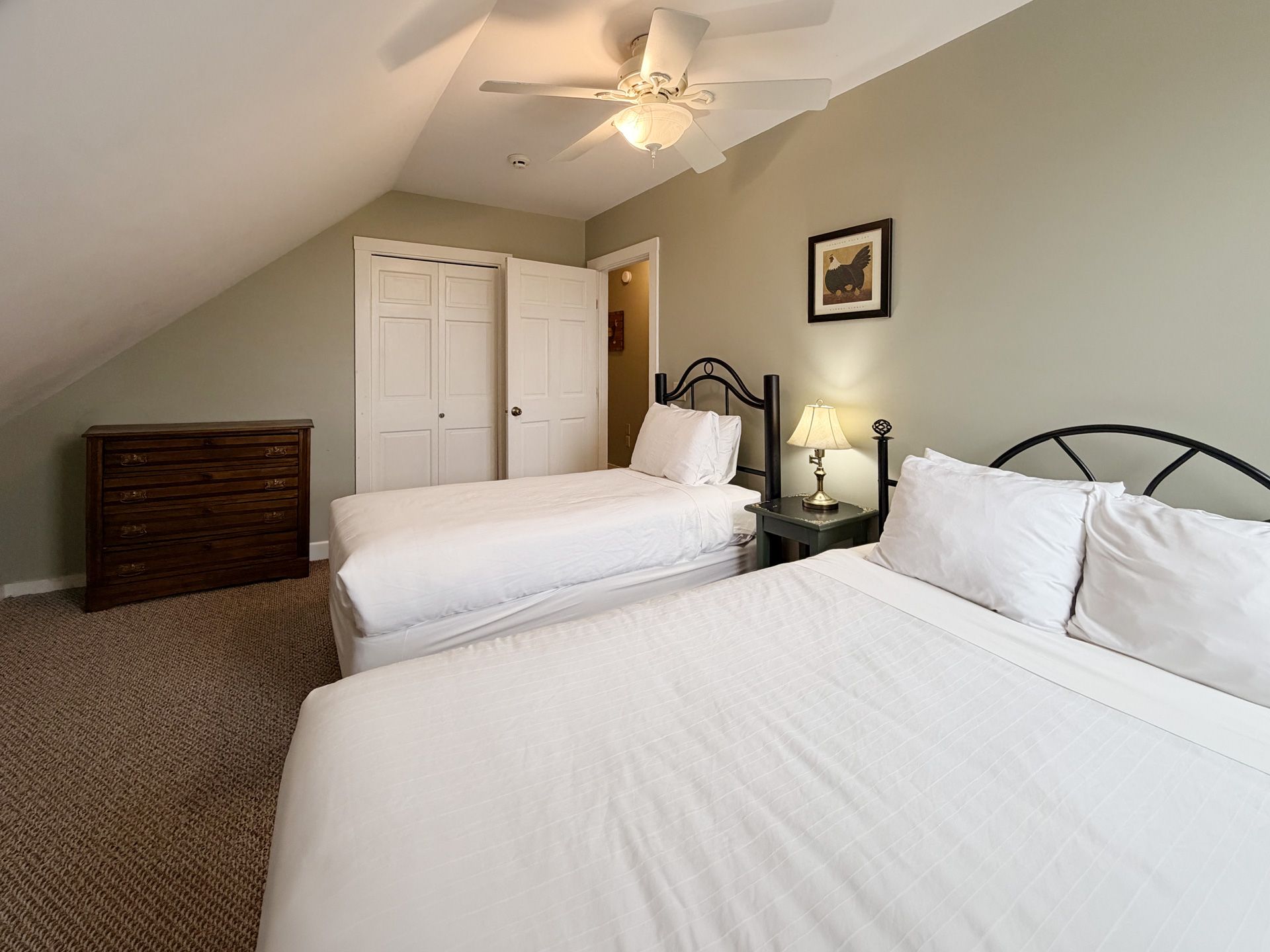 Bedroom with two white beds, sloped ceiling, ceiling fan, dresser, and framed wall art.