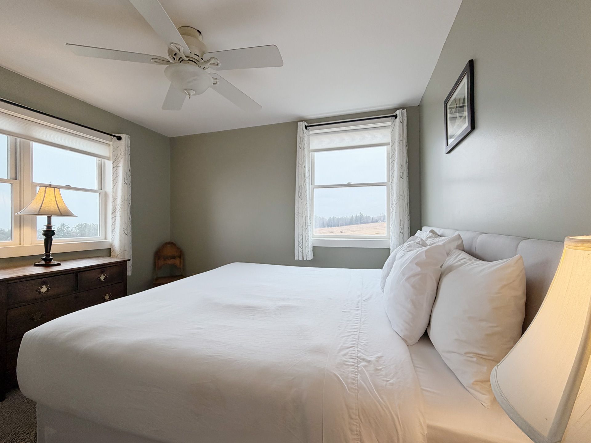 Bright bedroom with white bed, gray walls, windows, ceiling fan, and a wooden dresser with lamp.