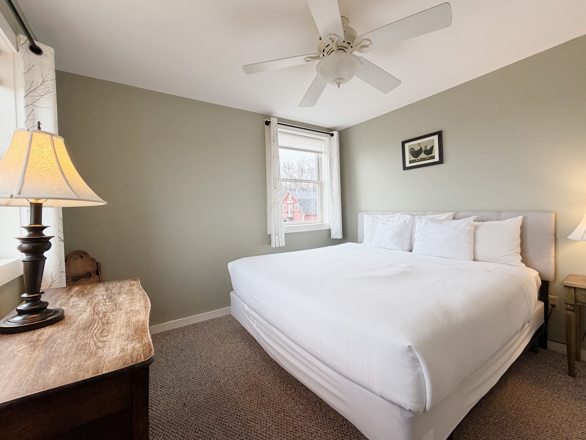 Bright bedroom with a made white bed, beige walls, ceiling fan, lamp, and window with a patterned shade.