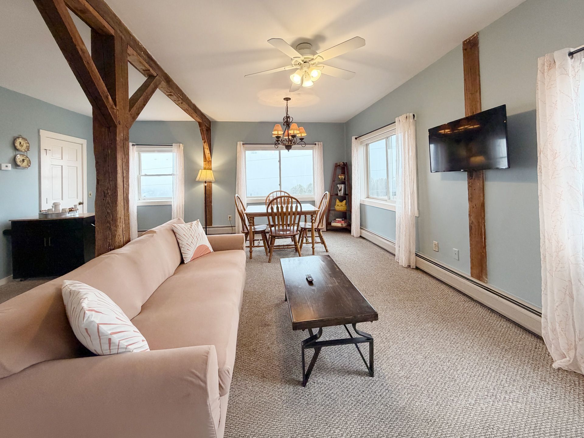Bright living-dining room with beige sofa, wooden beams, carpet, and wall-mounted TV