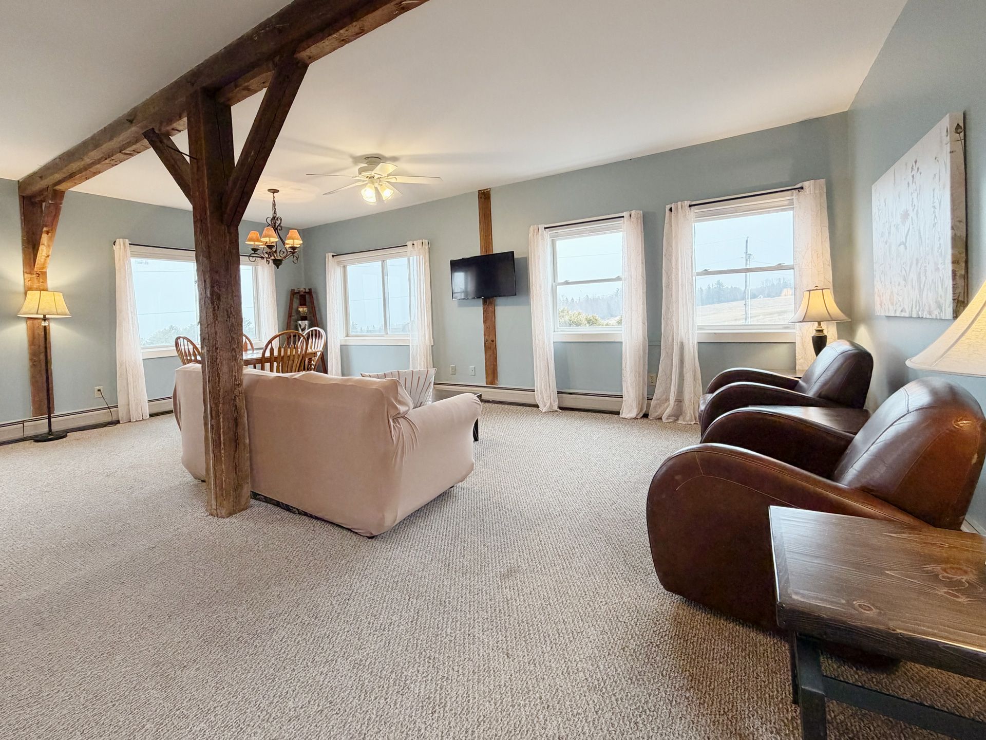 Spacious living room with beige carpet, wooden beams, leather chairs, and large windows overlooking water.