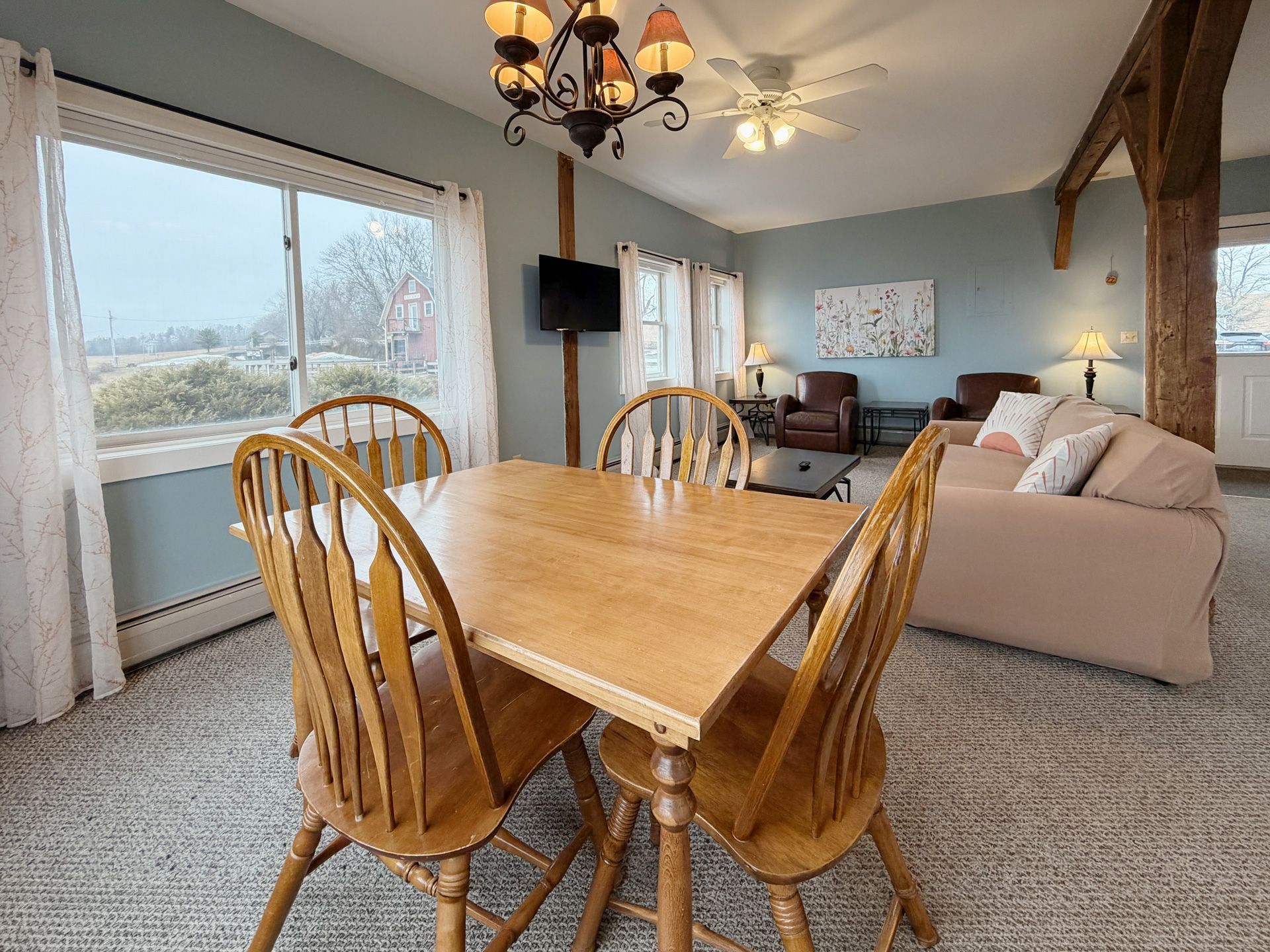 Bright open dining and living room with wood table, beige sofa, wall-mounted TV, and large windows.