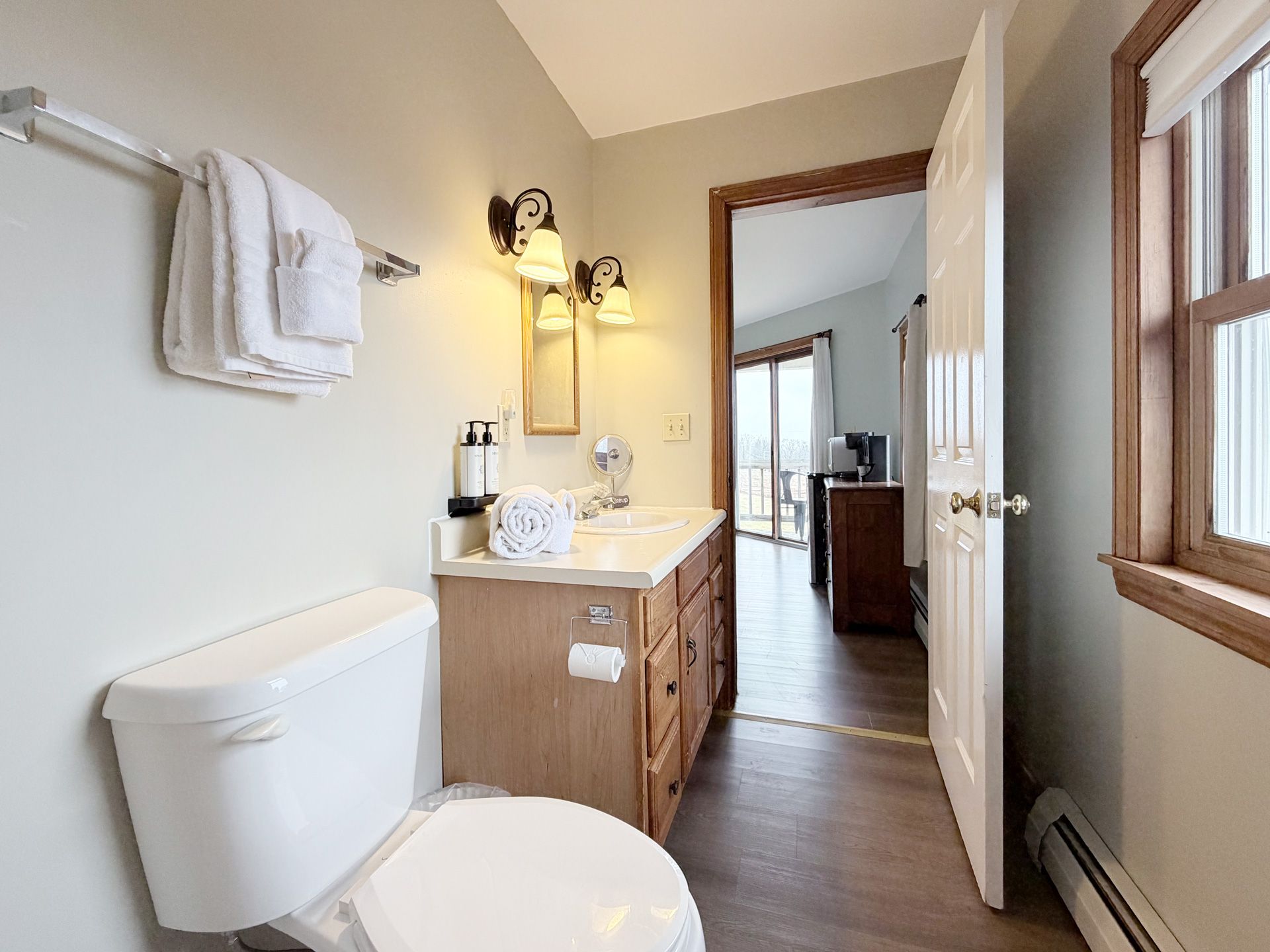 Small bathroom with white toilet, wood vanity, sink, mirror, and open door to a bedroom.