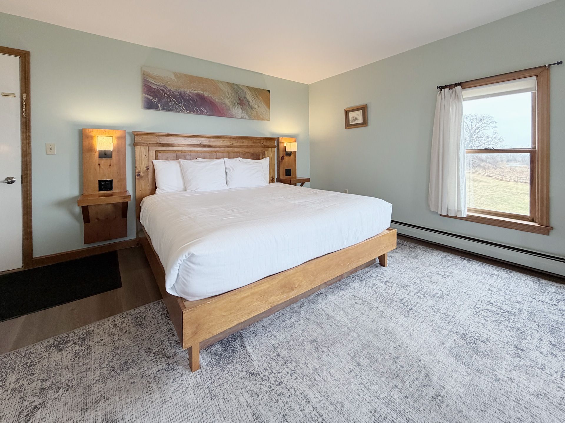 Bright bedroom with a wooden bed, white bedding, gray carpet, and a window with sheer curtains.
