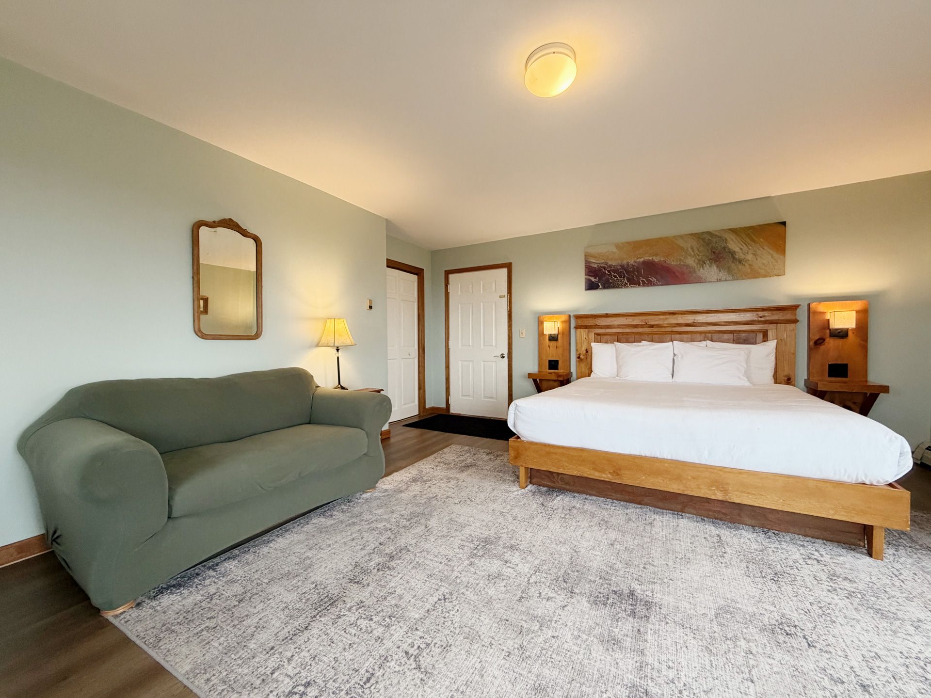 Spacious bedroom with a bed, green sofa, lamp, mirror, and light carpeted floor.