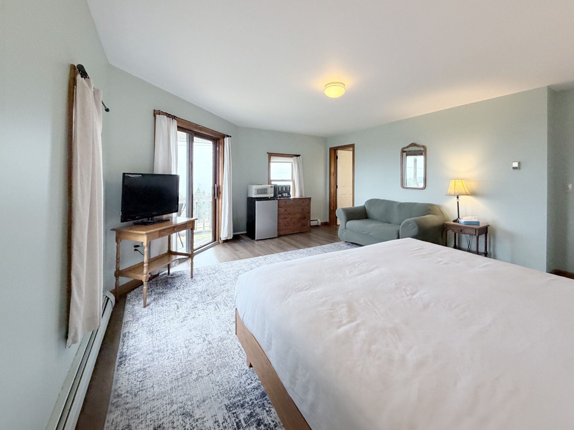 Bright bedroom with a white bed, gray sofa, TV, and windows letting in natural light.