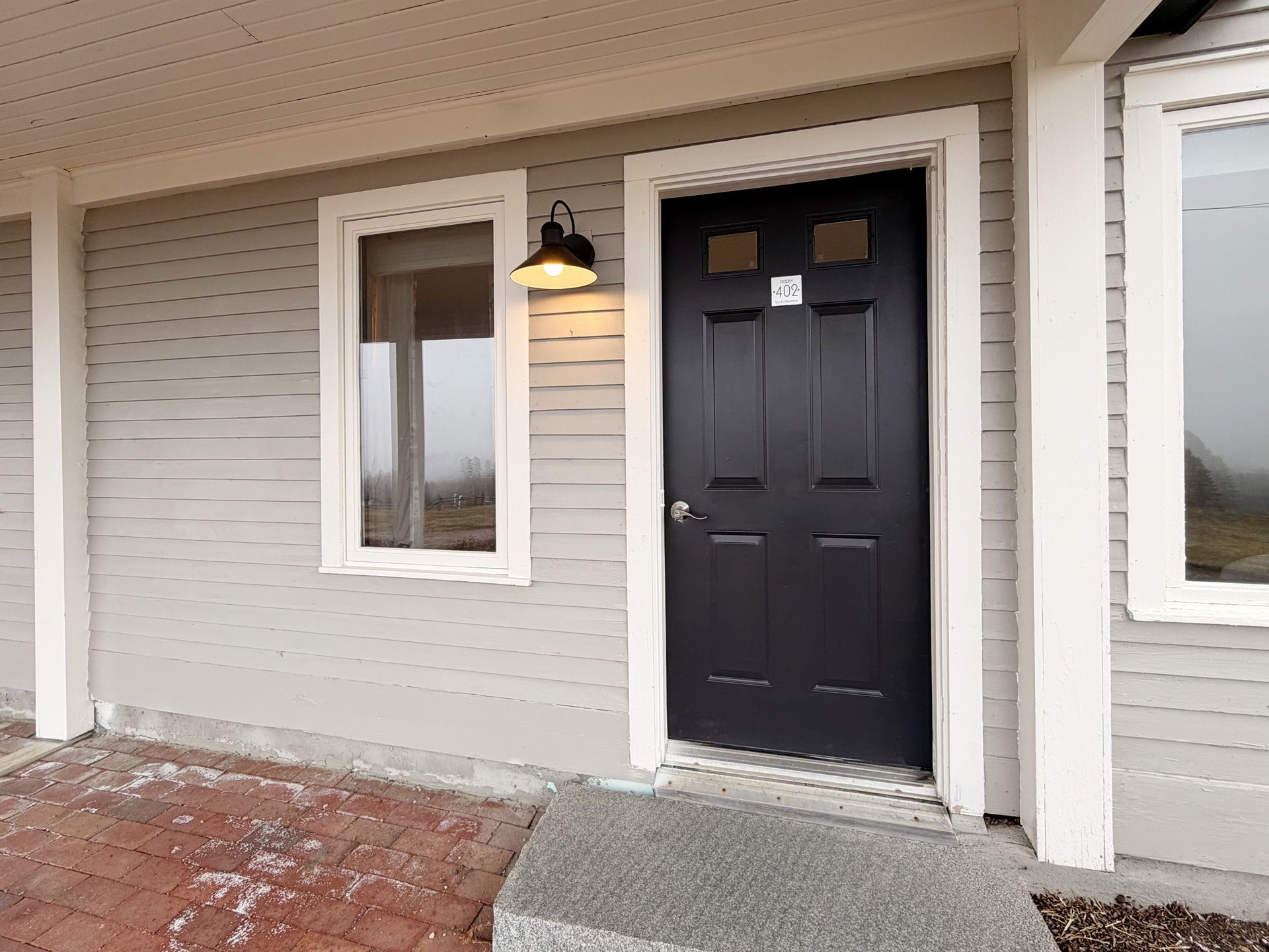 Front porch with a black door, wall light, and two windows on a brick-paved entry.