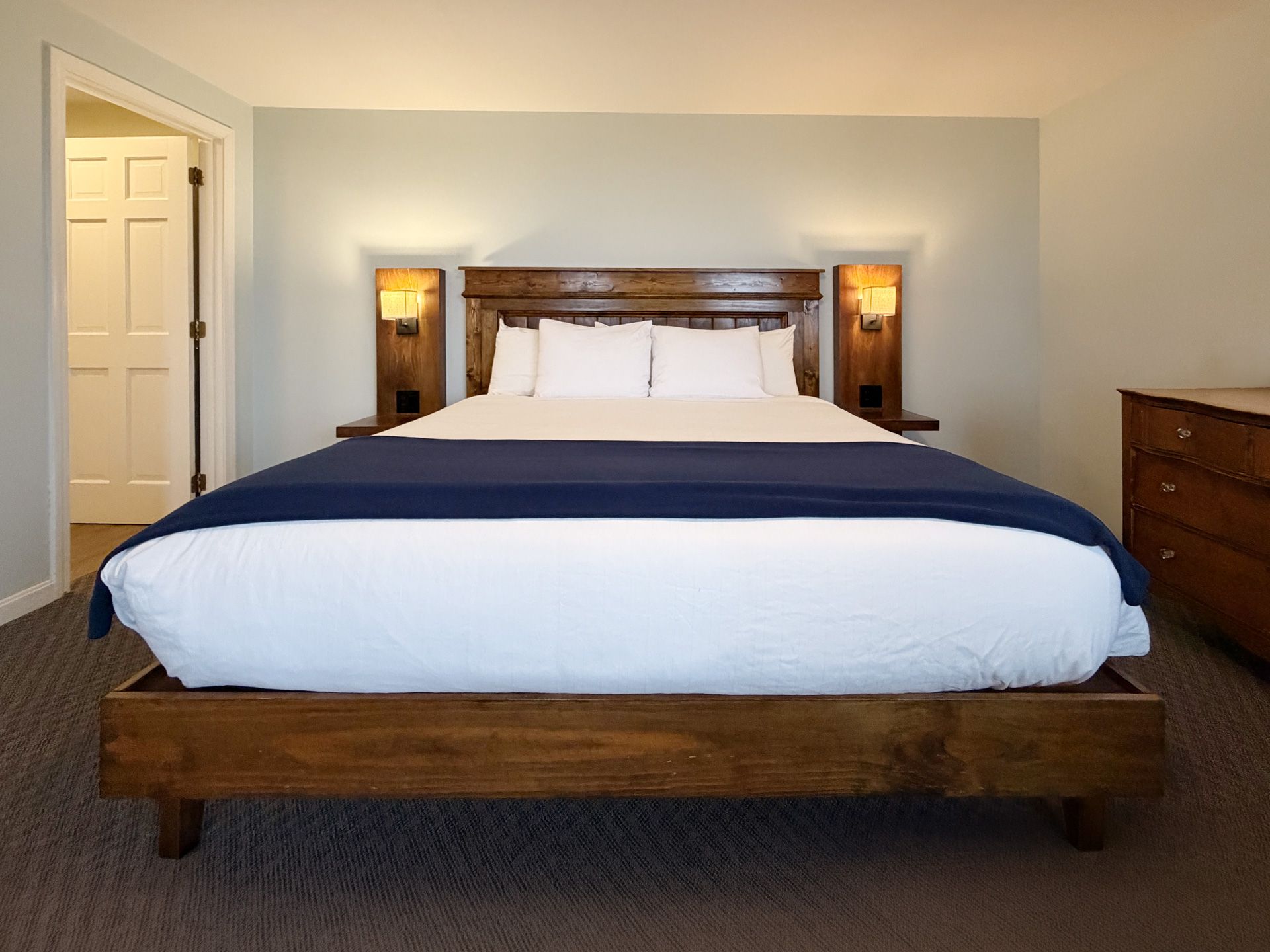 Cozy bedroom with a wooden bed, navy coverlet, lit lamps, and dresser