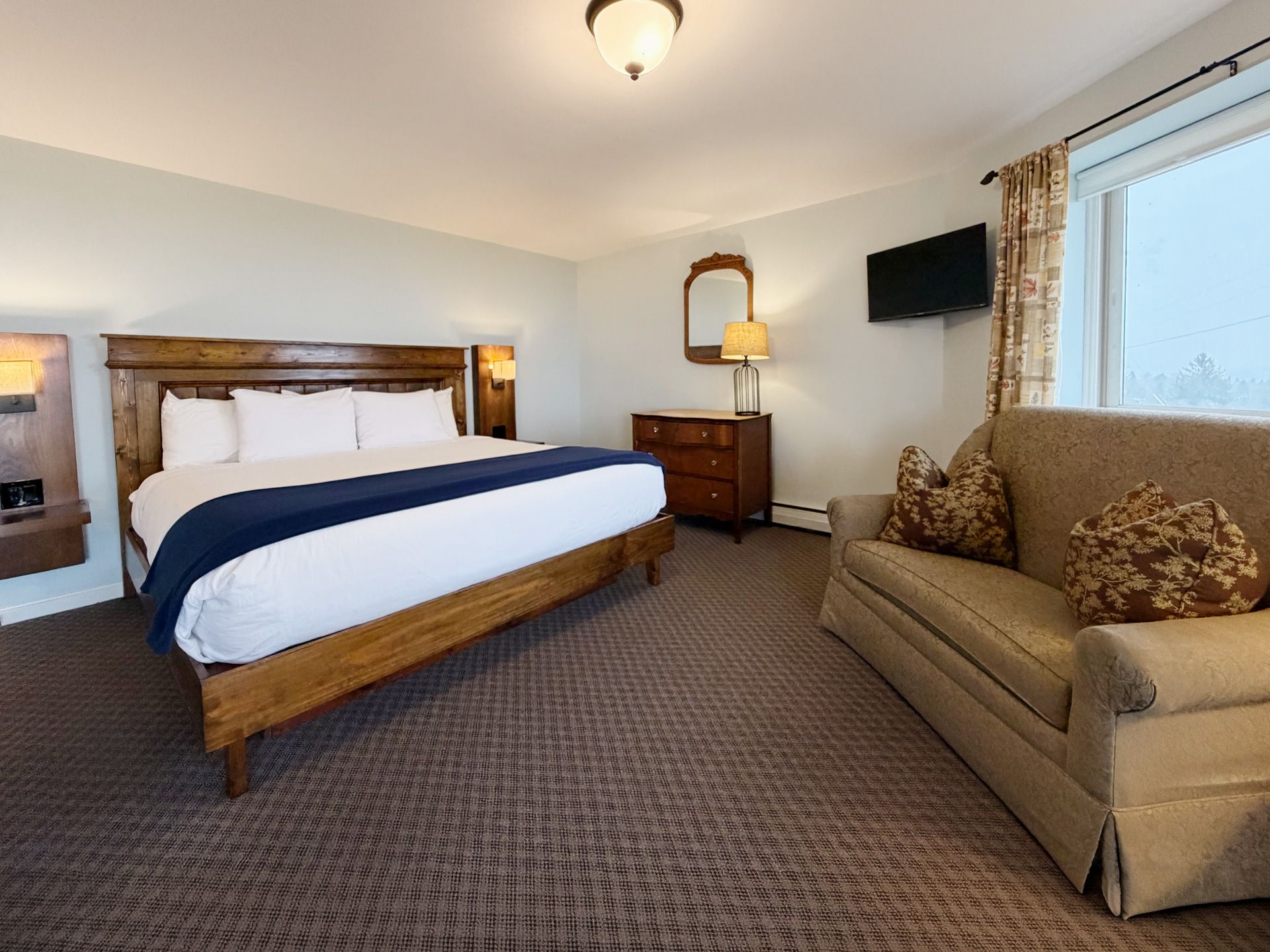 Hotel bedroom with a king bed, tan sofa, wall TV, and patterned carpet.