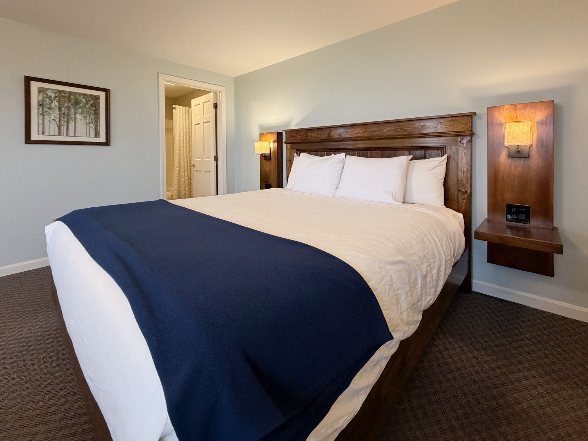 Hotel room with a neatly made bed, navy runner, wooden headboard, and bedside lamps