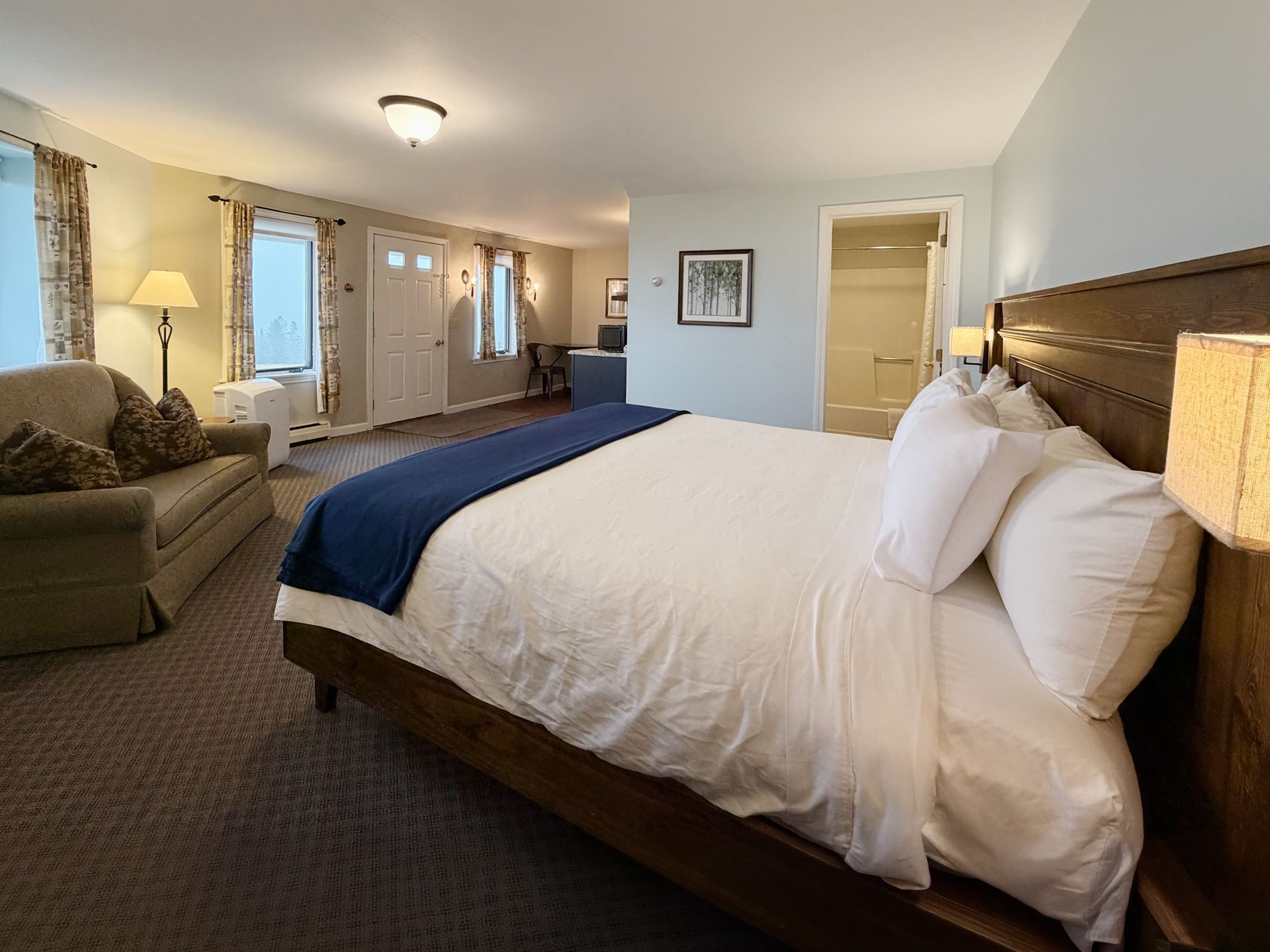 Hotel bedroom with white bed, blue runner, armchair, desk, and lamps in a softly lit room