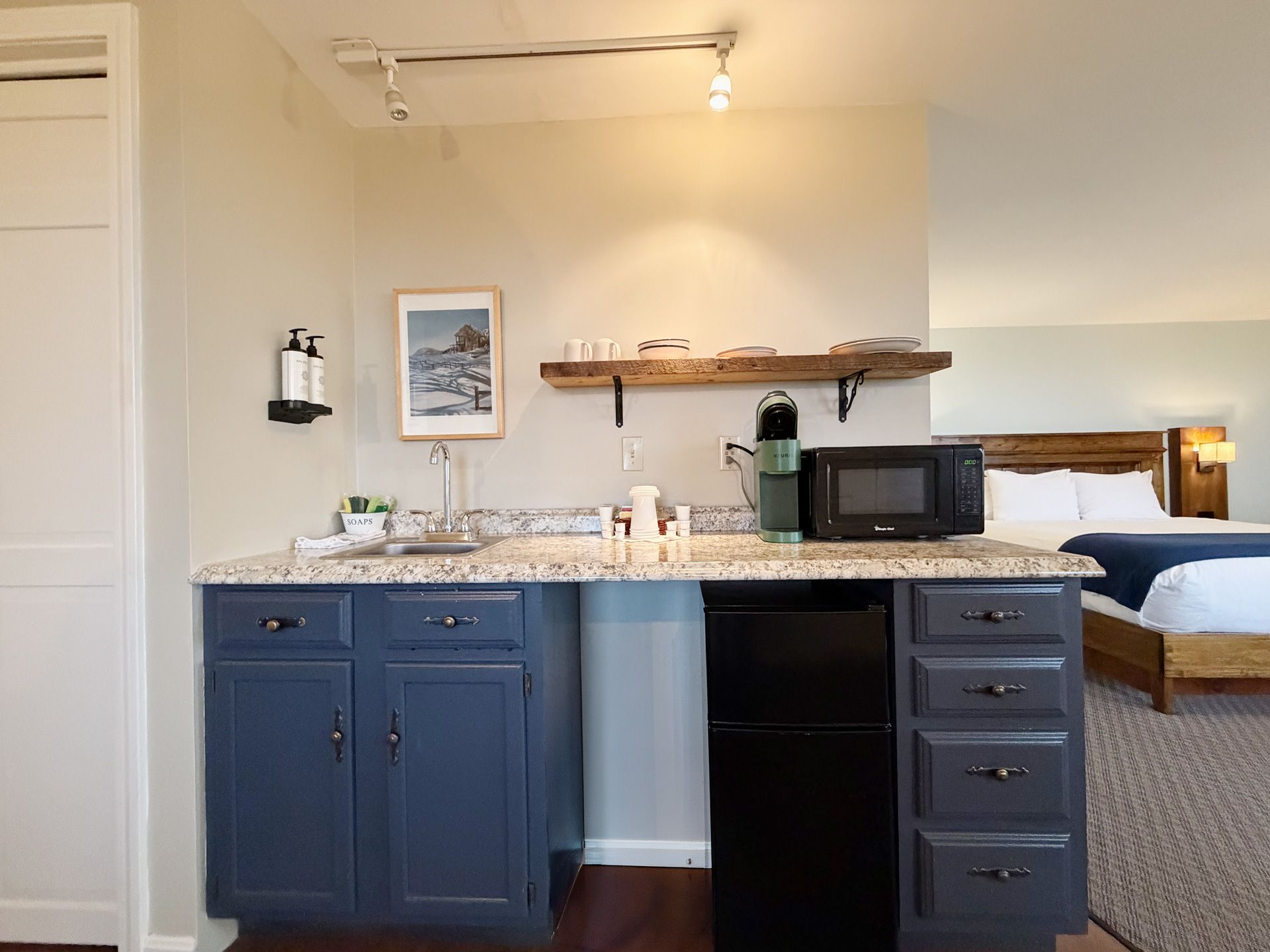 Compact kitchenette with blue cabinets, granite counter, microwave, and a bed in the adjoining room