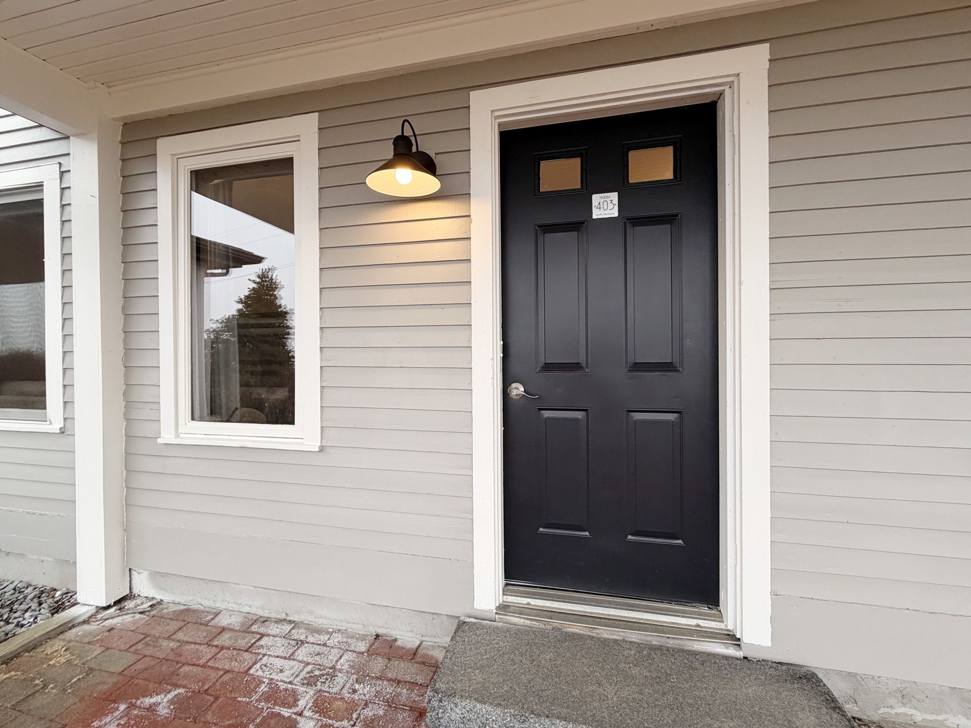 Snow-dusted porch with a black front door and white siding beside a window