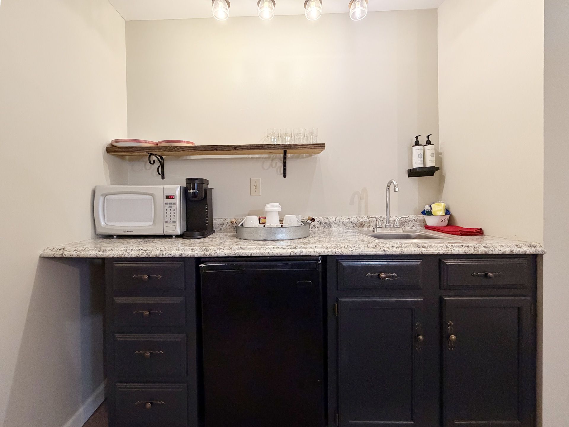 Small kitchenette with dark cabinets, speckled countertop, microwave, coffee maker, and wall shelf.