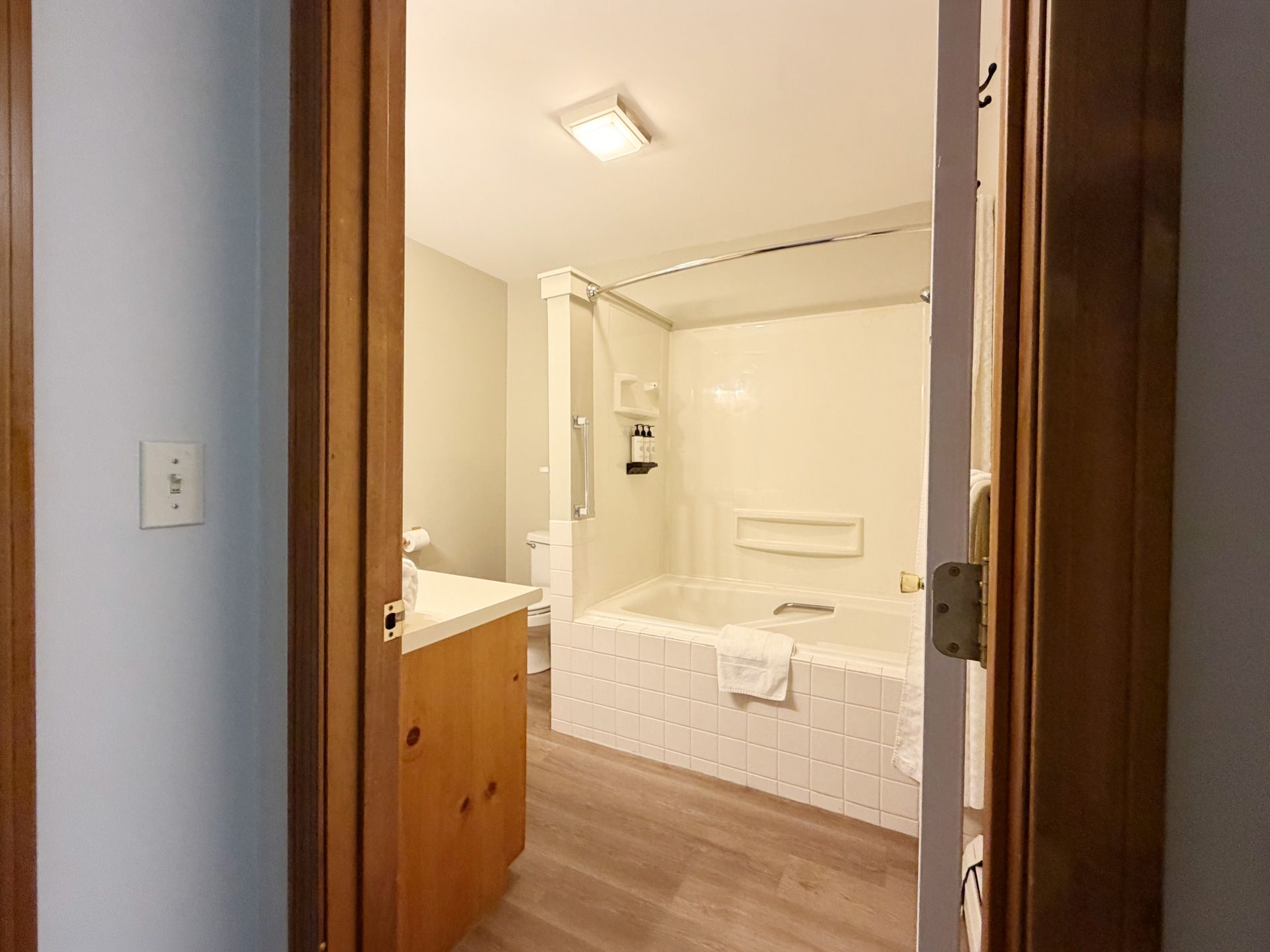 Bathroom with white tub-shower, wood vanity, and tiled floor, viewed through a doorway