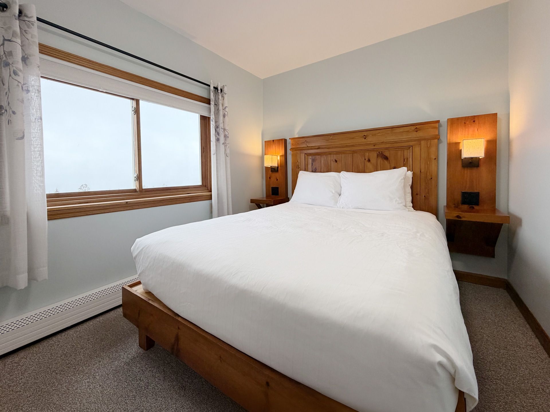 Bright bedroom with a wooden bed, white bedding, bedside lamps, and a window with curtains.