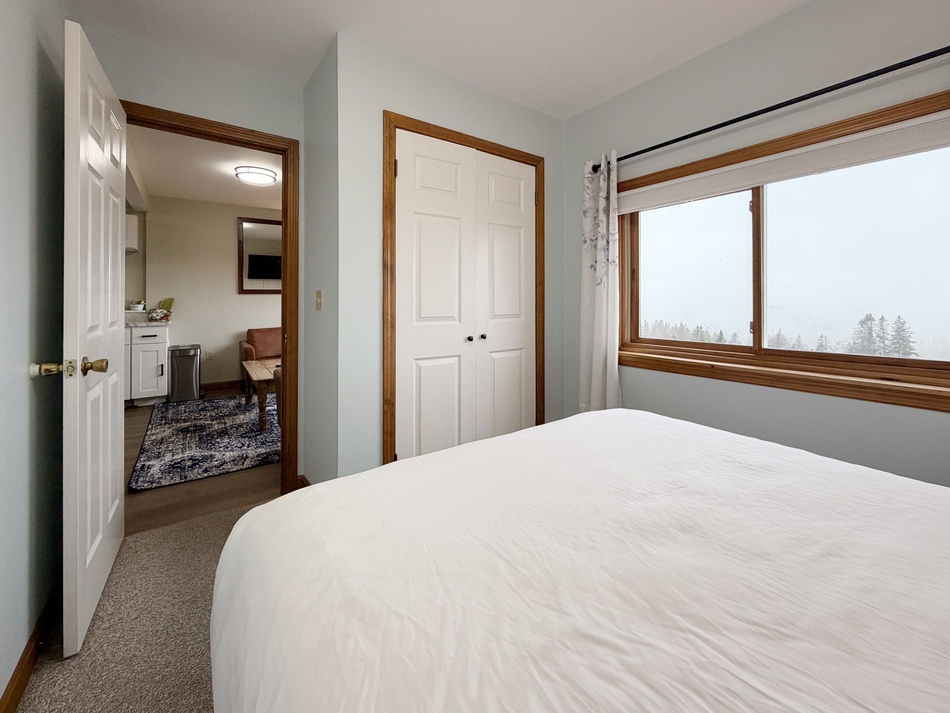 Bright bedroom with a white bed, open door, and large window showing a snowy view.