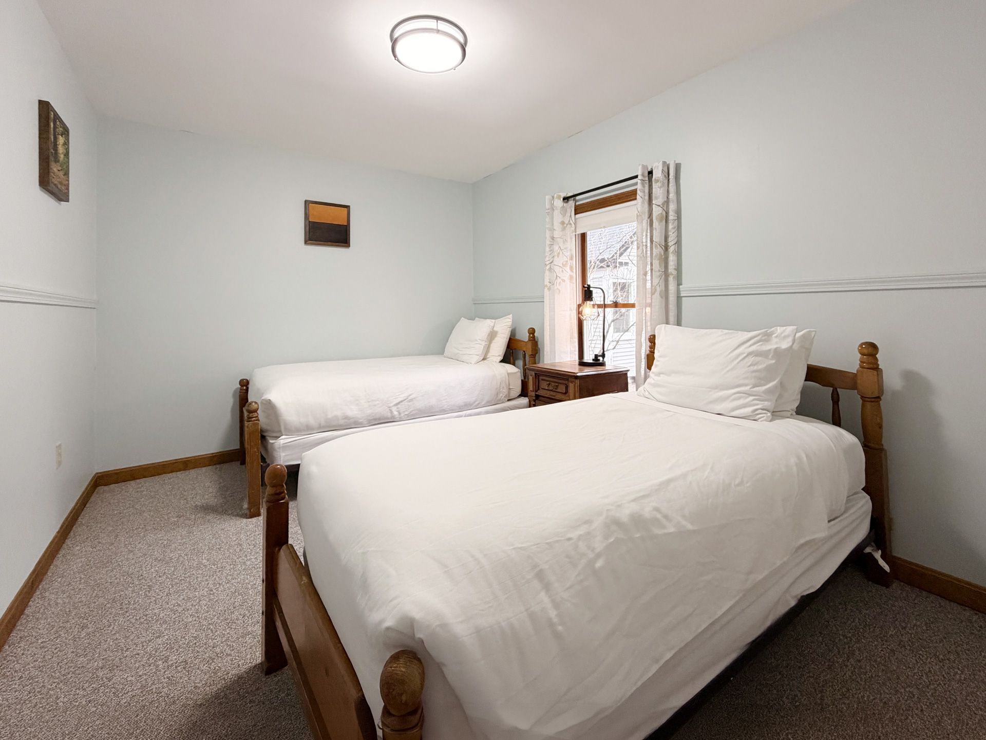 Small bedroom with two white beds, pale walls, and a window with blinds and curtains
