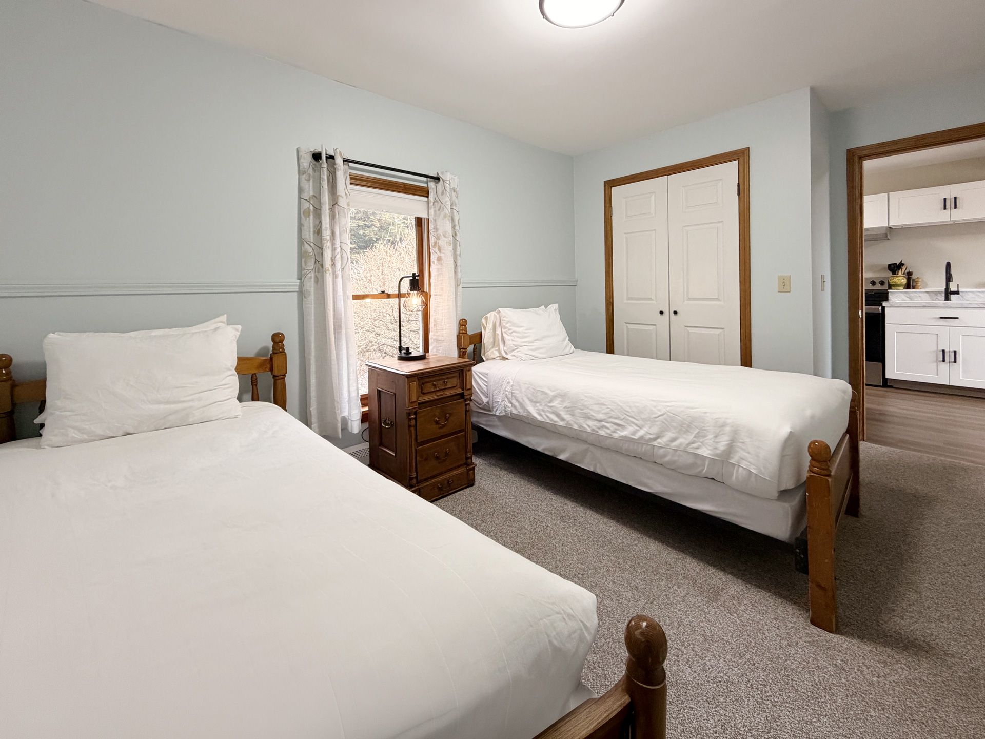 Bright bedroom with two white beds, a wooden dresser, mirrored closet, and open doorway to a bathroom.