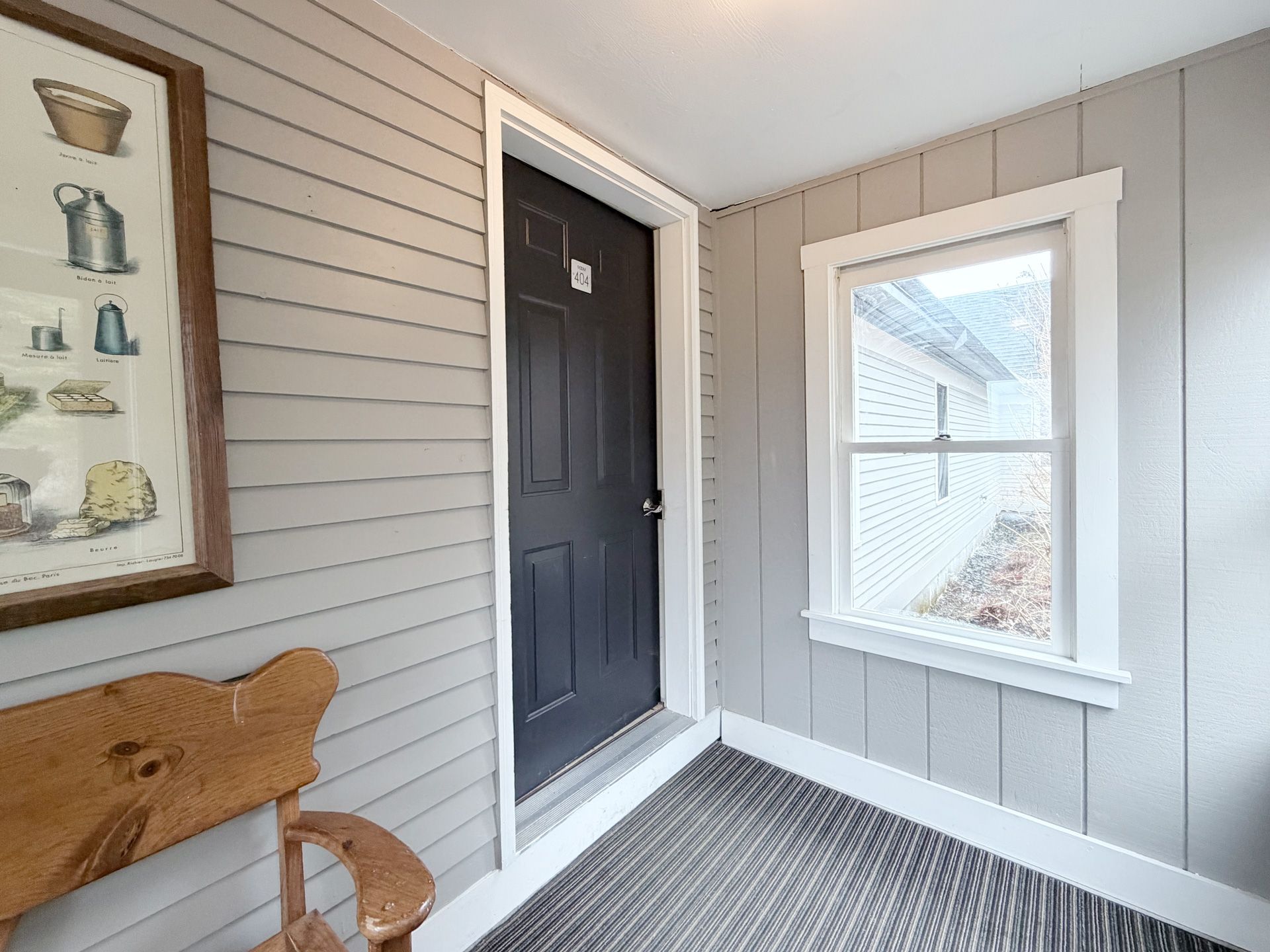 Front porch with gray siding, black door, window, and a wooden bench with wall art.
