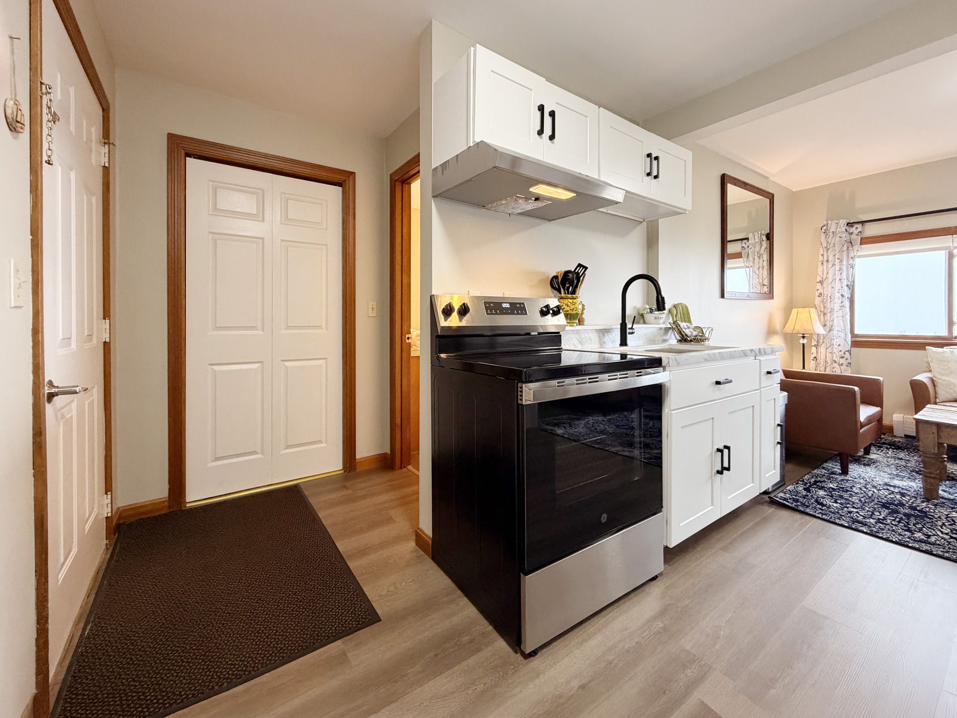 Hallway view into a small kitchen with black stove, white cabinets, and a cozy living room beyond.