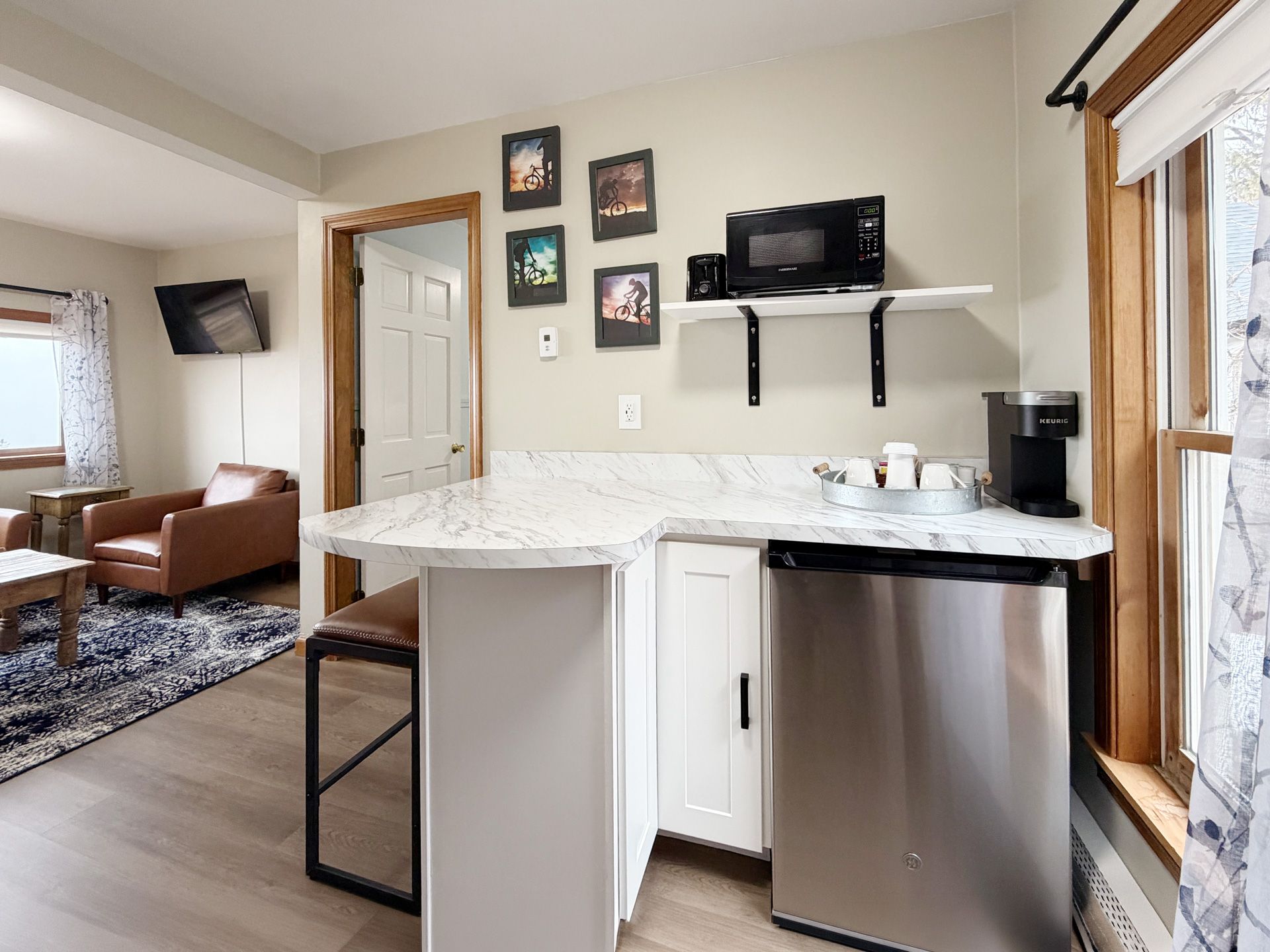 Bright kitchen nook with white island, stainless mini fridge, shelves, and a view into a cozy living room.