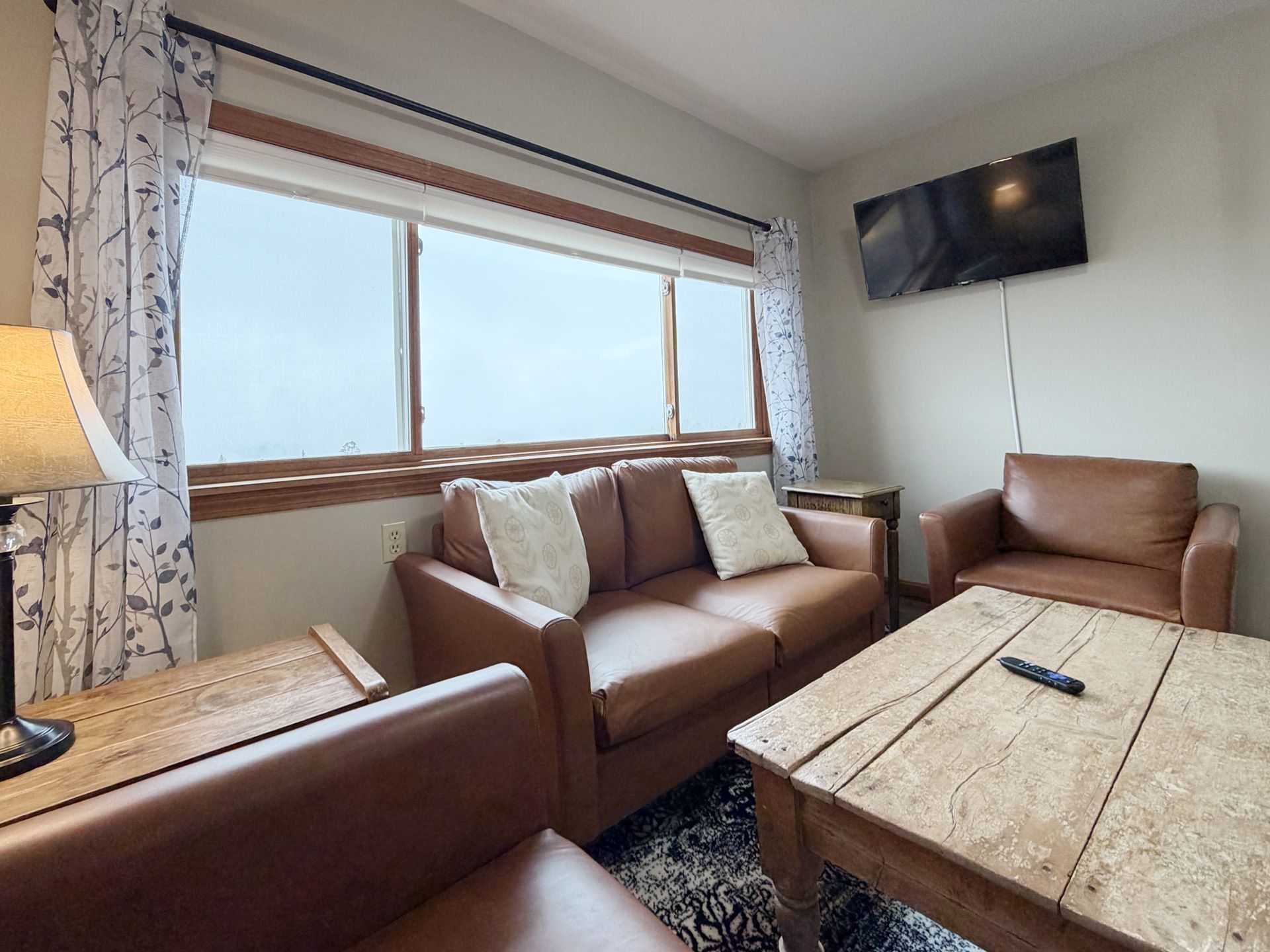 Cozy living room with brown leather sofas, rustic coffee table, large window, and wall-mounted TV