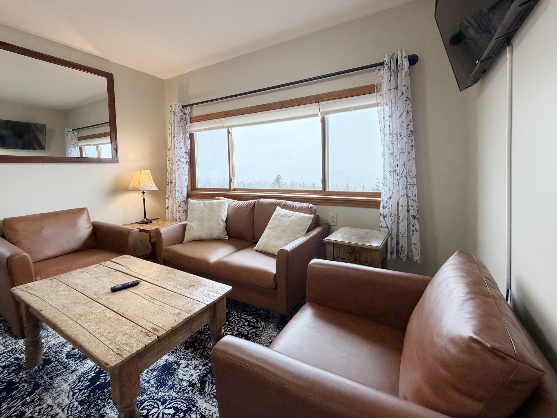 Cozy living room with brown leather sofas, rustic coffee table, lamp, mirror, and large window with curtains