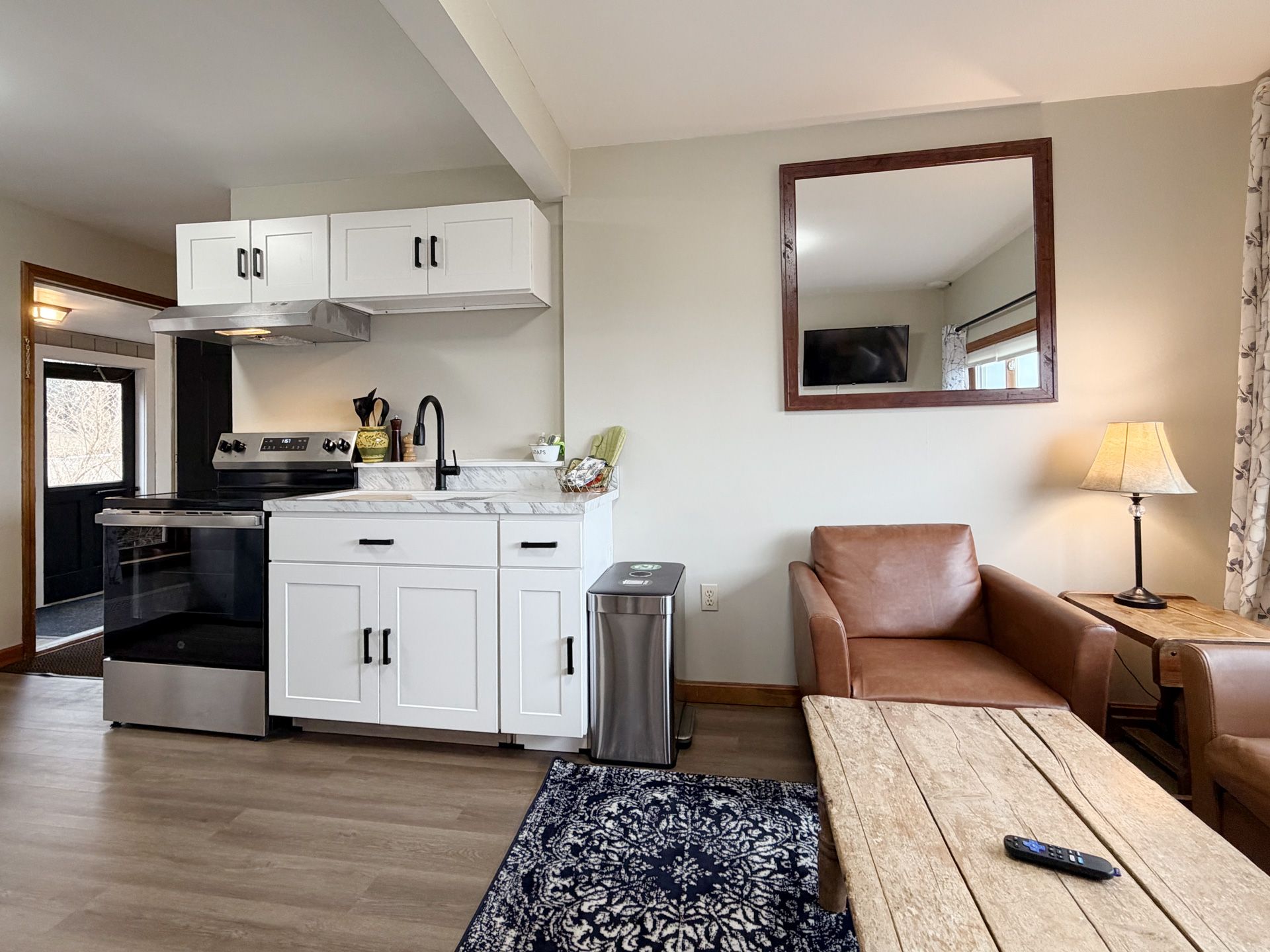 Small living room with kitchenette, brown leather chair, wall mirror, and lit table lamp.